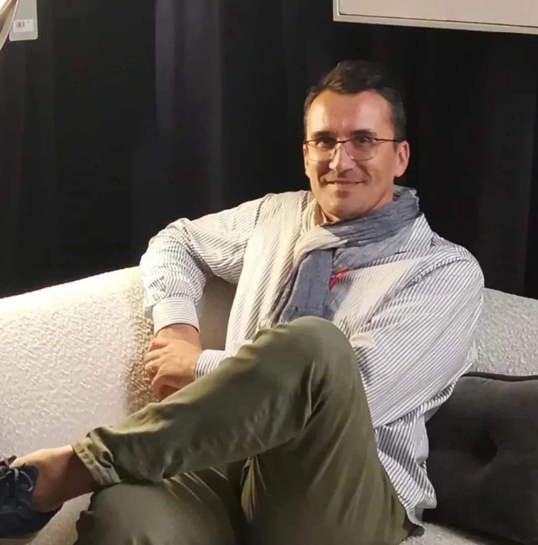 Homme souriant assis sur un canapé, portant des lunettes, une chemise à rayures, une écharpe grise et des pantalons kaki, dans un intérieur sombre.