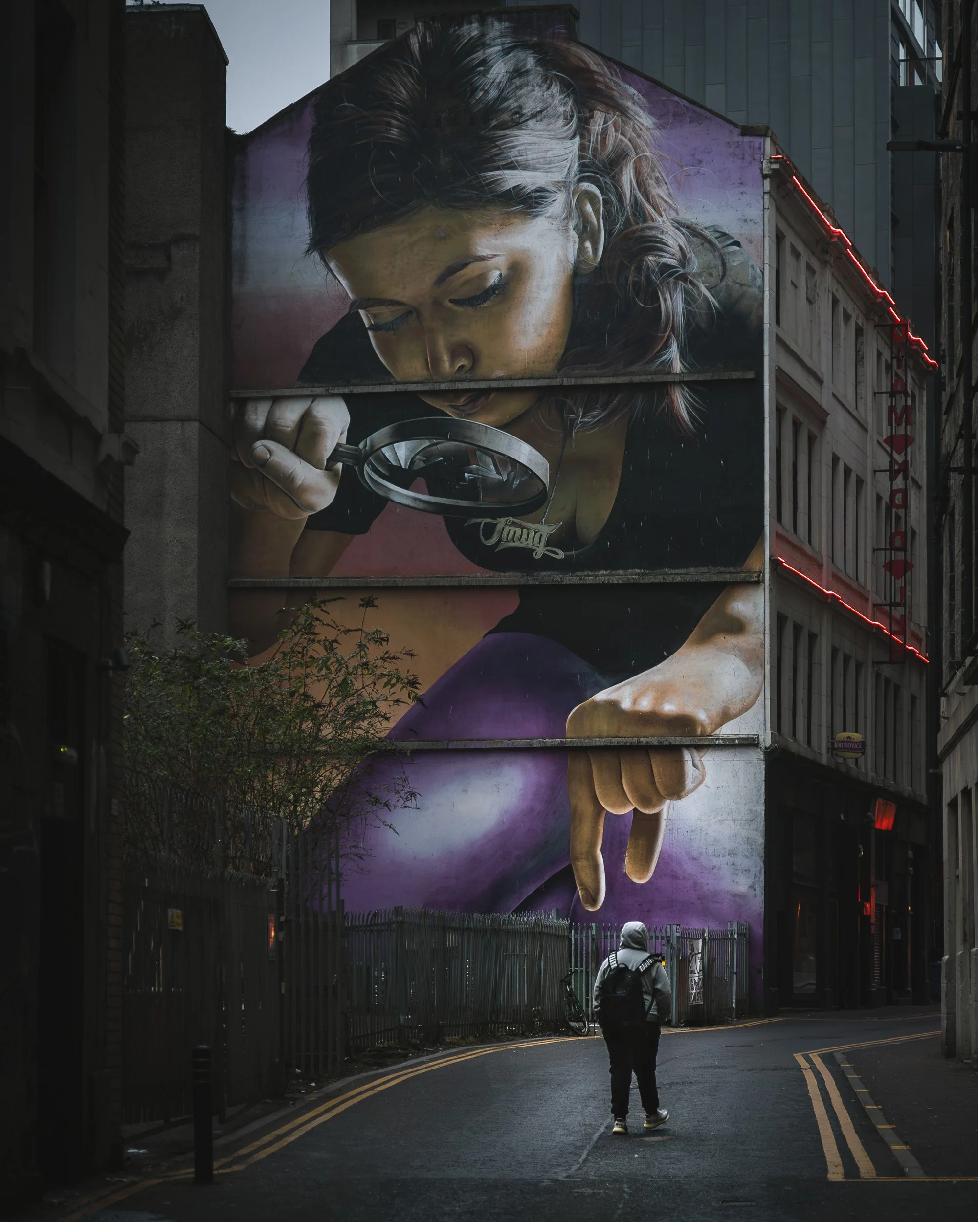 A hooded man walks beneath a giant mural of a woman holding a magnifying glass in Glasgow, aligned with her painted fingers above the street.