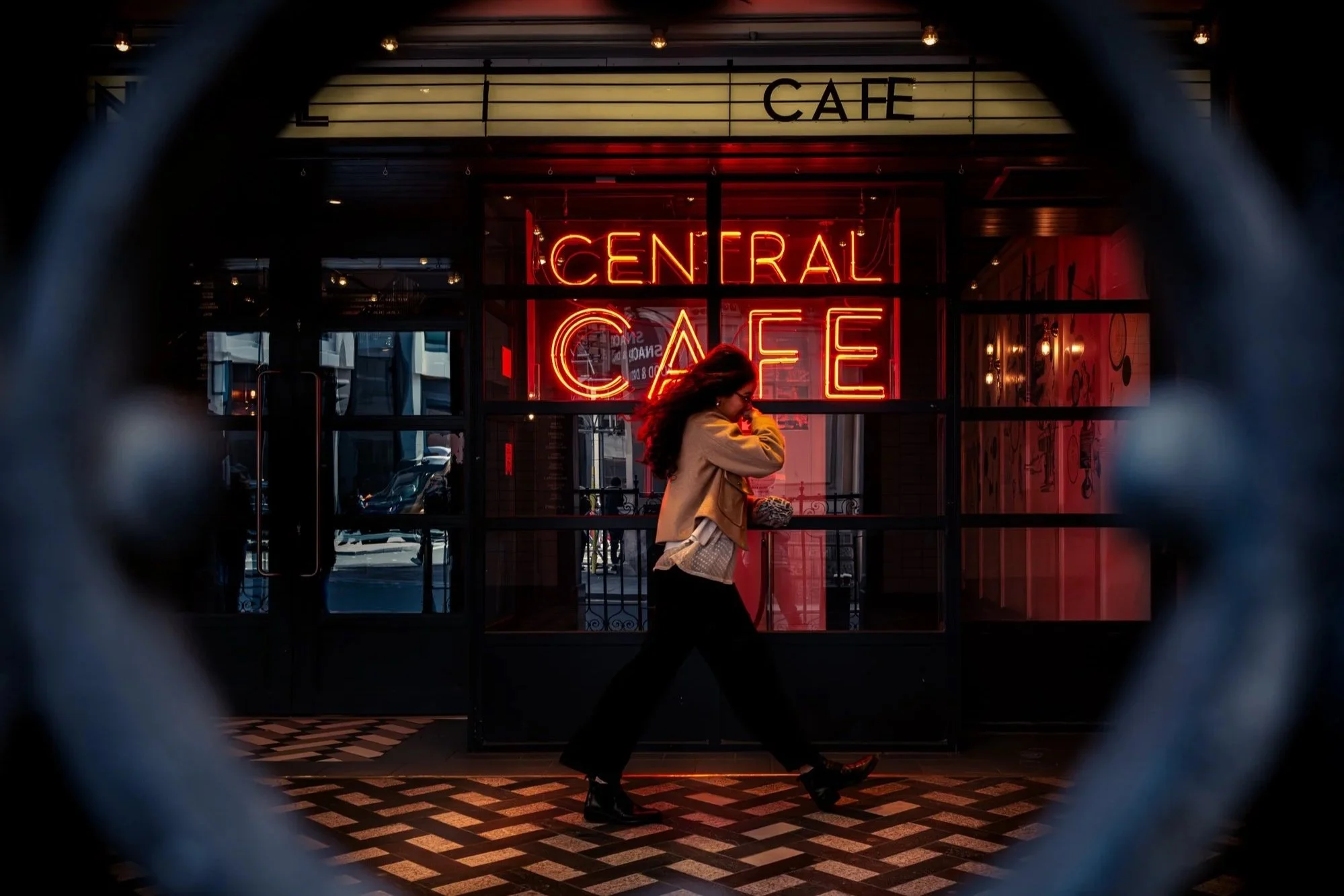 A woman walking past a cafe with a neon sign that reads 'Central Cafe' in red letters. The cafe has glass doors and windows, with reflections and a street scene visible outside. The woman has long hair, is wearing a beige jacket, black pants, and black shoes, and is carrying a small purse.