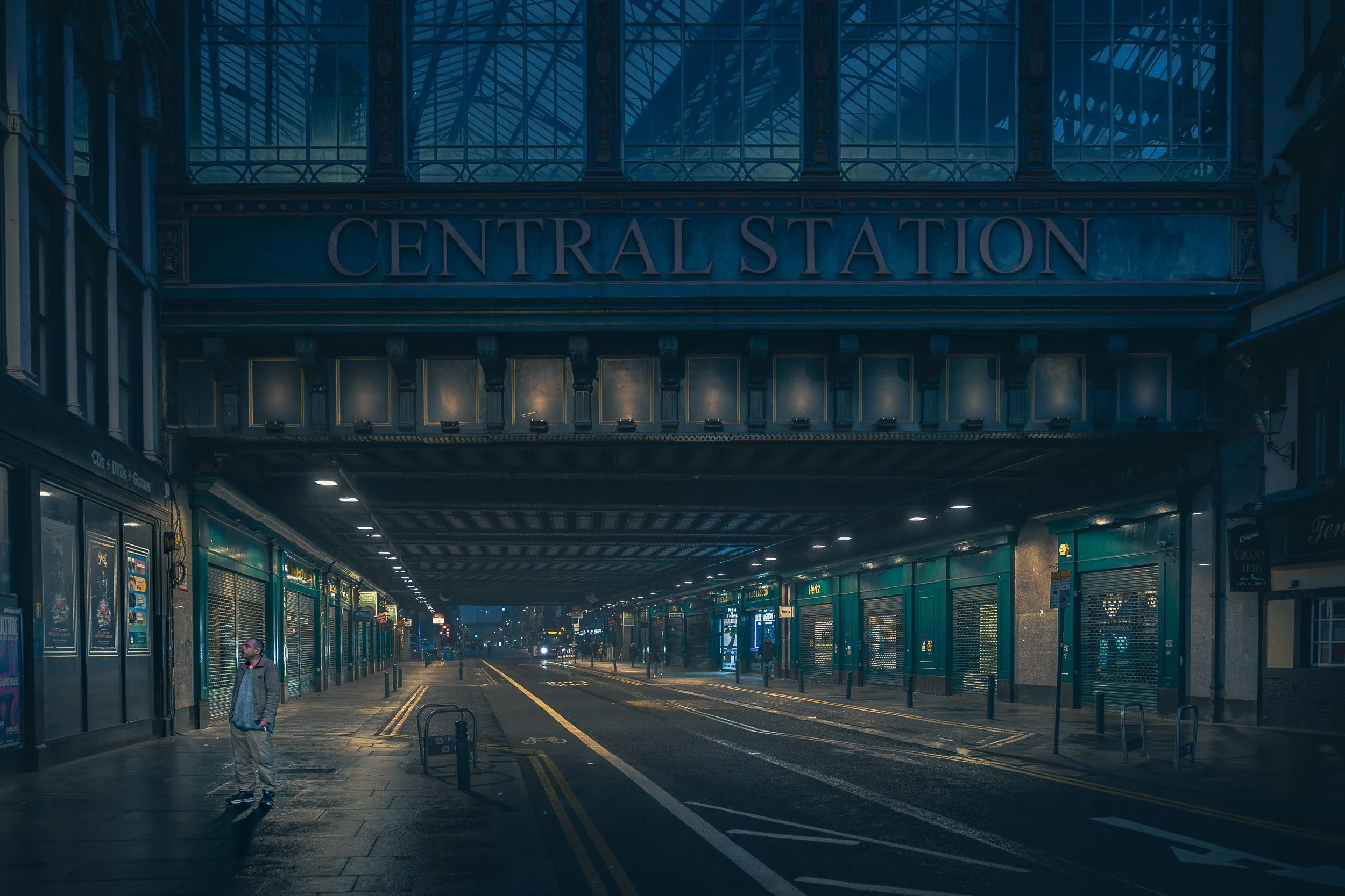 A man stands beneath Glasgow Central Station in the early morning, surrounded by blue light, shuttered shops and warm pools of street lighting.
