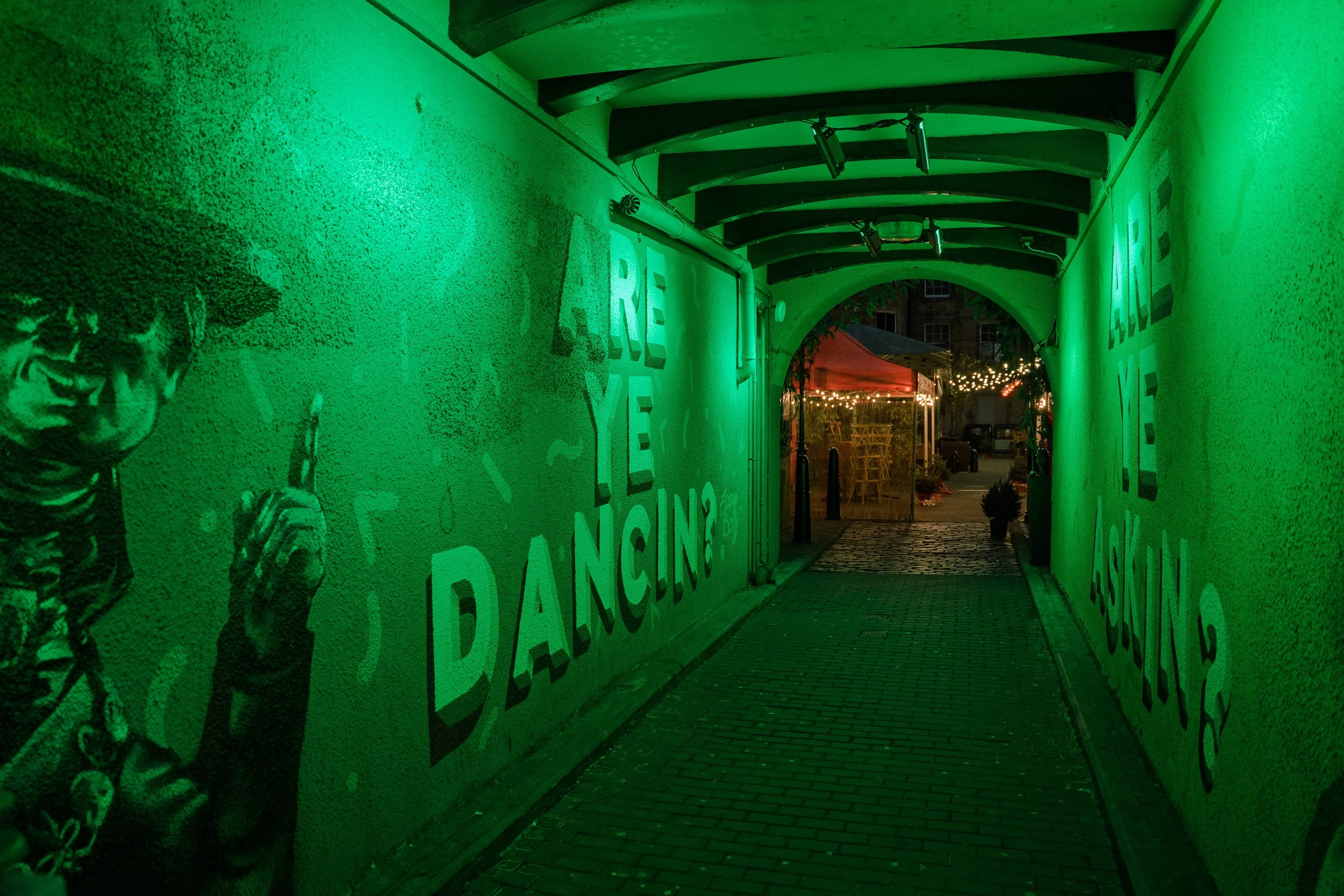 Green-lit passageway with murals leading towards a warmly lit courtyard in Glasgow.