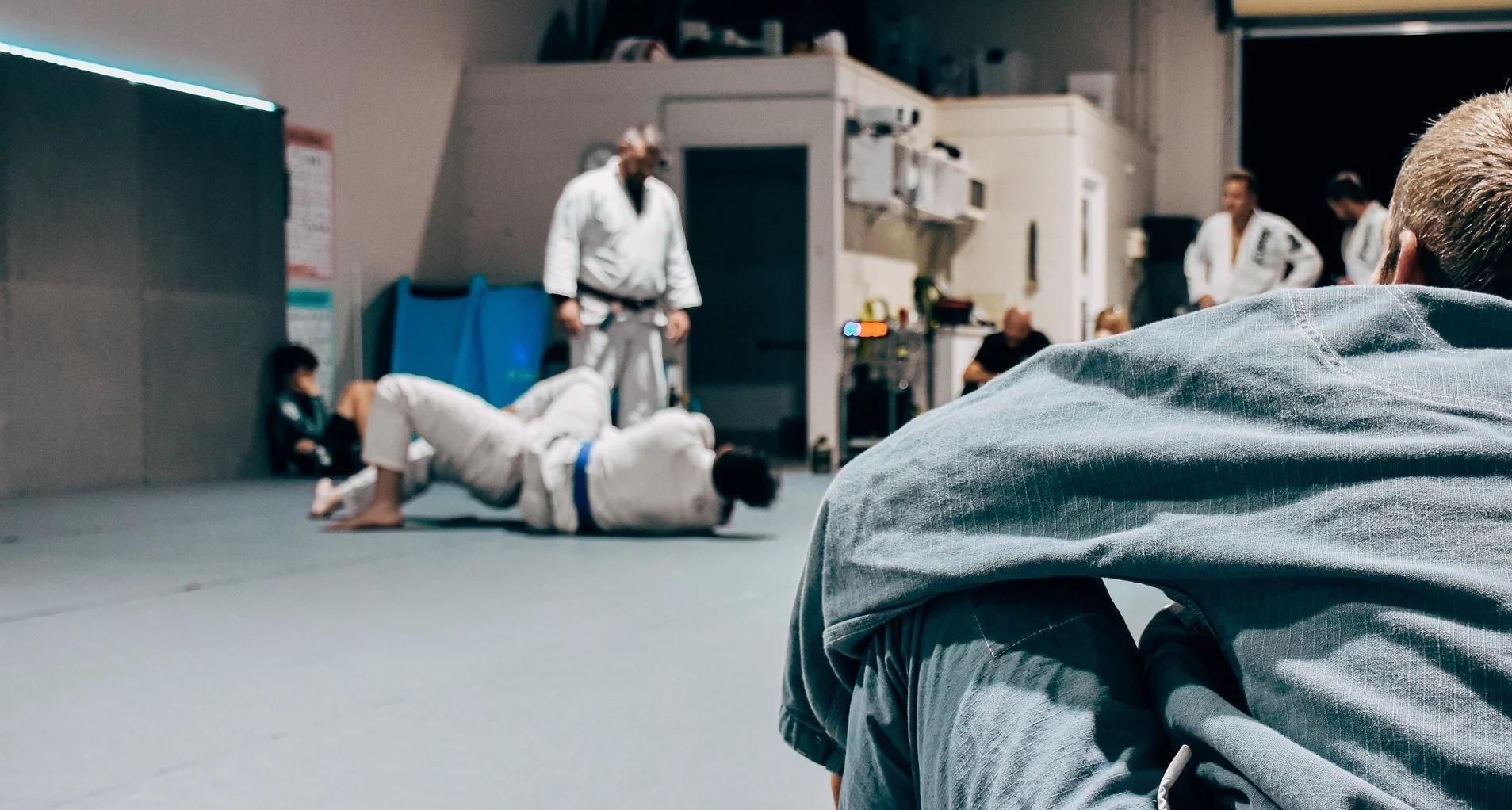 Martial arts training in a dojo with multiple students. One student is on the ground practicing grappling, while another is observing. Several people are seated or standing, watching the practice. The room has a gray padded floor and fitness equipment along the walls.