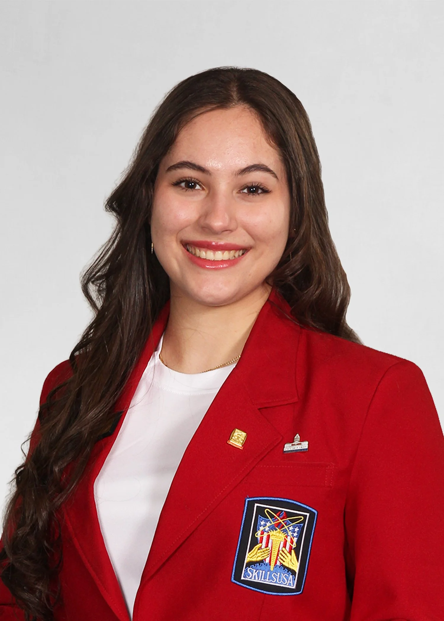 A young woman smiling outdoors in front of a cityscape with buildings and trees, wearing a blue SkillsUSA Massachusetts jacket with her arms crossed.