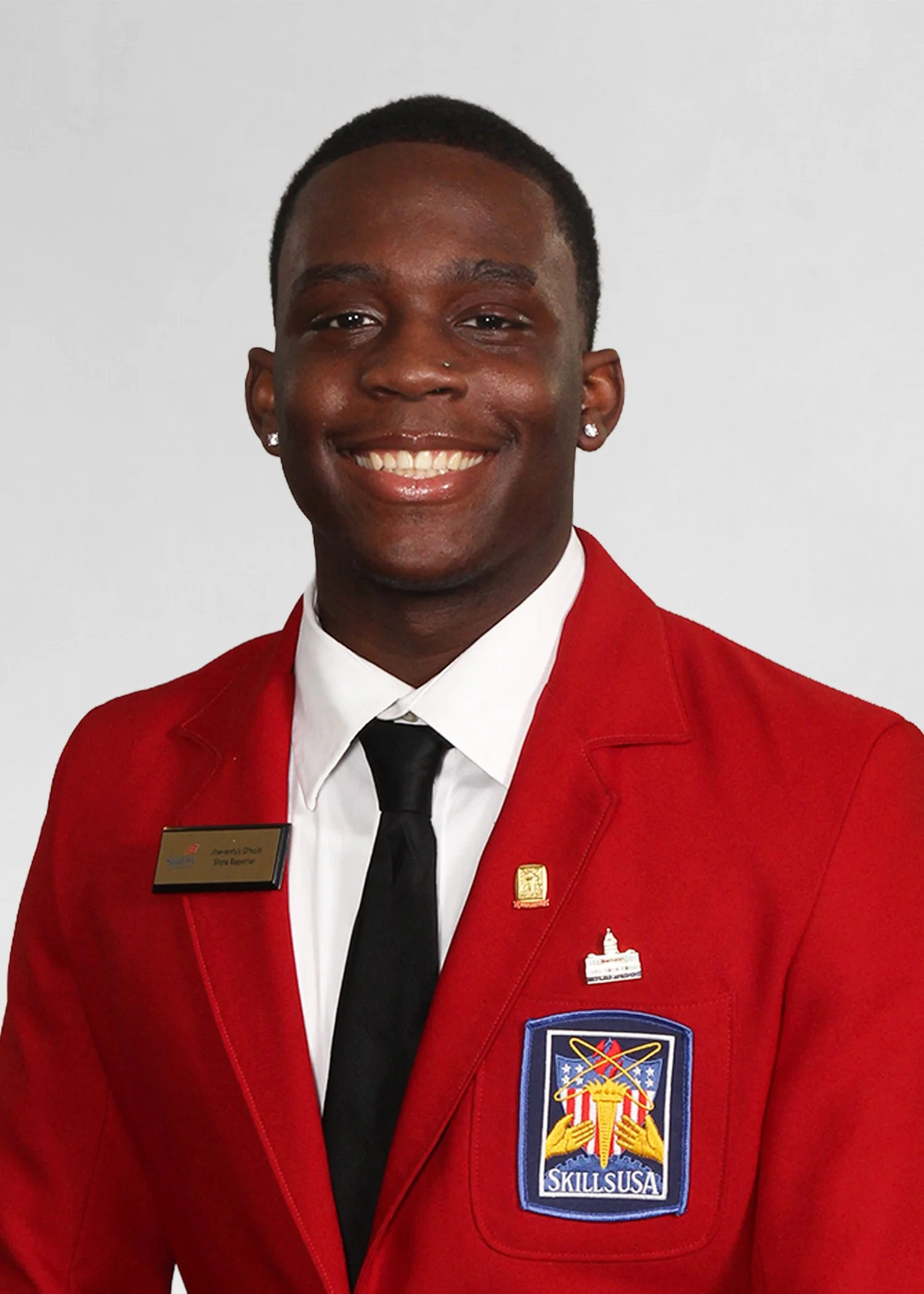 Close-up portrait of a young African American man with styled hair, wearing earrings, a navy blue jacket with 'STATE OFFICER' and 'SkillsUSA MASSACHUSETTS' embroidered, outdoors with trees and buildings in the background.