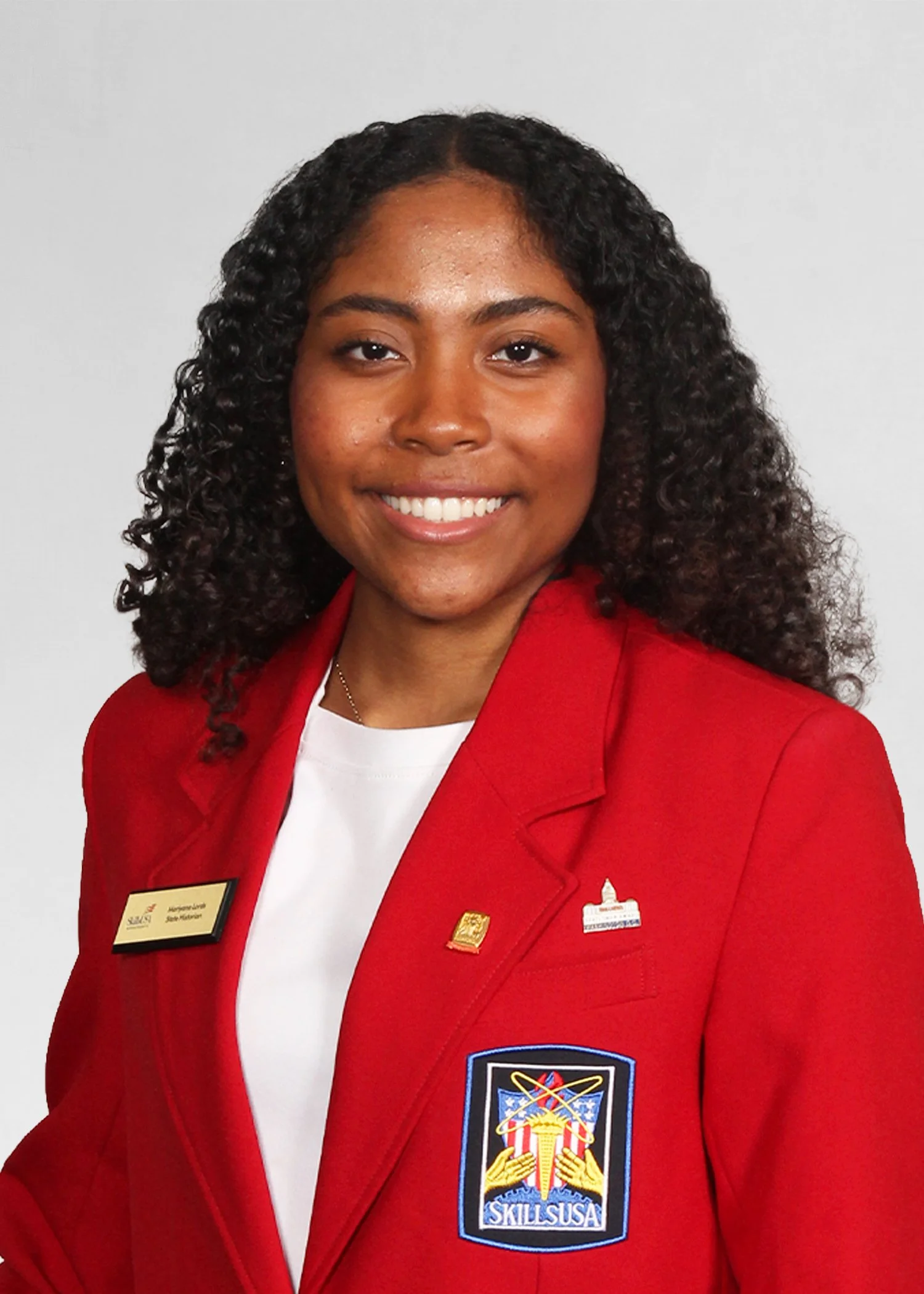 A young woman with curly hair smiling outdoors, wearing a navy blue SkillsUSA Massachusetts jacket and gold hoop earrings.