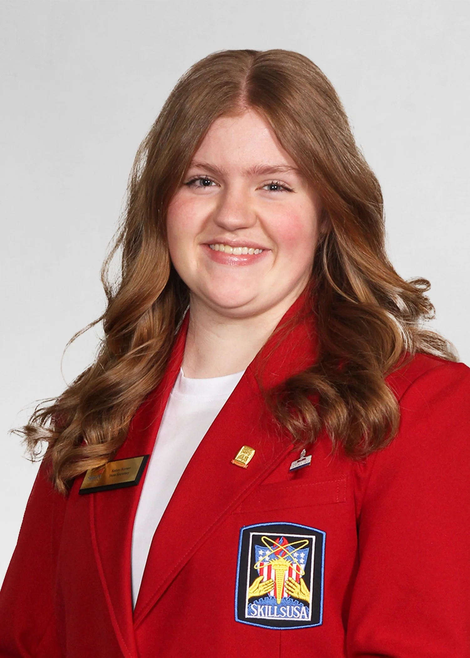 A young woman with long, wavy brown hair, wearing a navy blue zip-up jacket with white embroidery that reads 'State Officer' and 'SkillsUSA Massachusetts,' standing outdoors in front of buildings and trees, smiling with arms crossed.