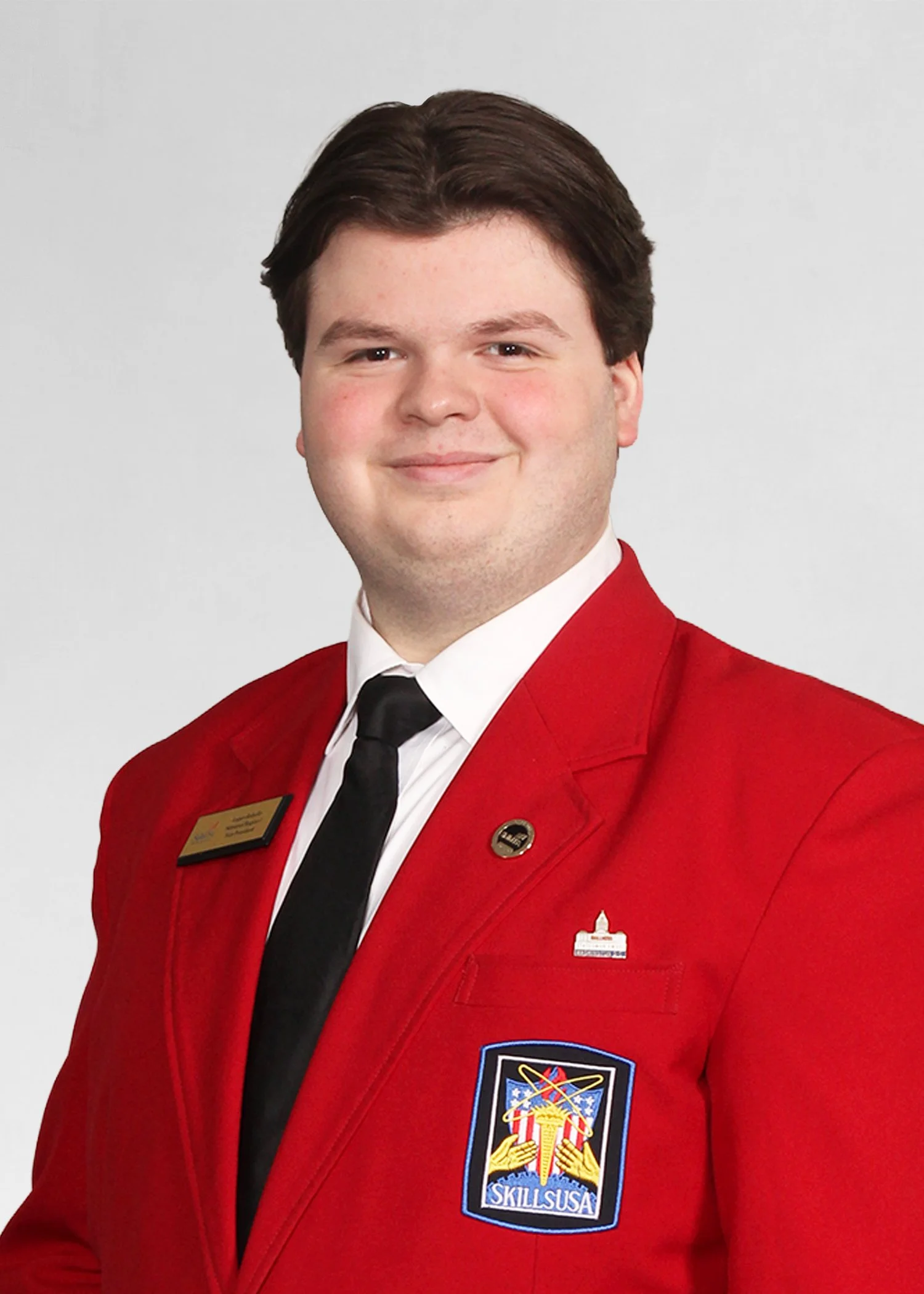 Young man smiling outdoors in urban setting, wearing a navy SkillsUSA Massachusetts jacket.