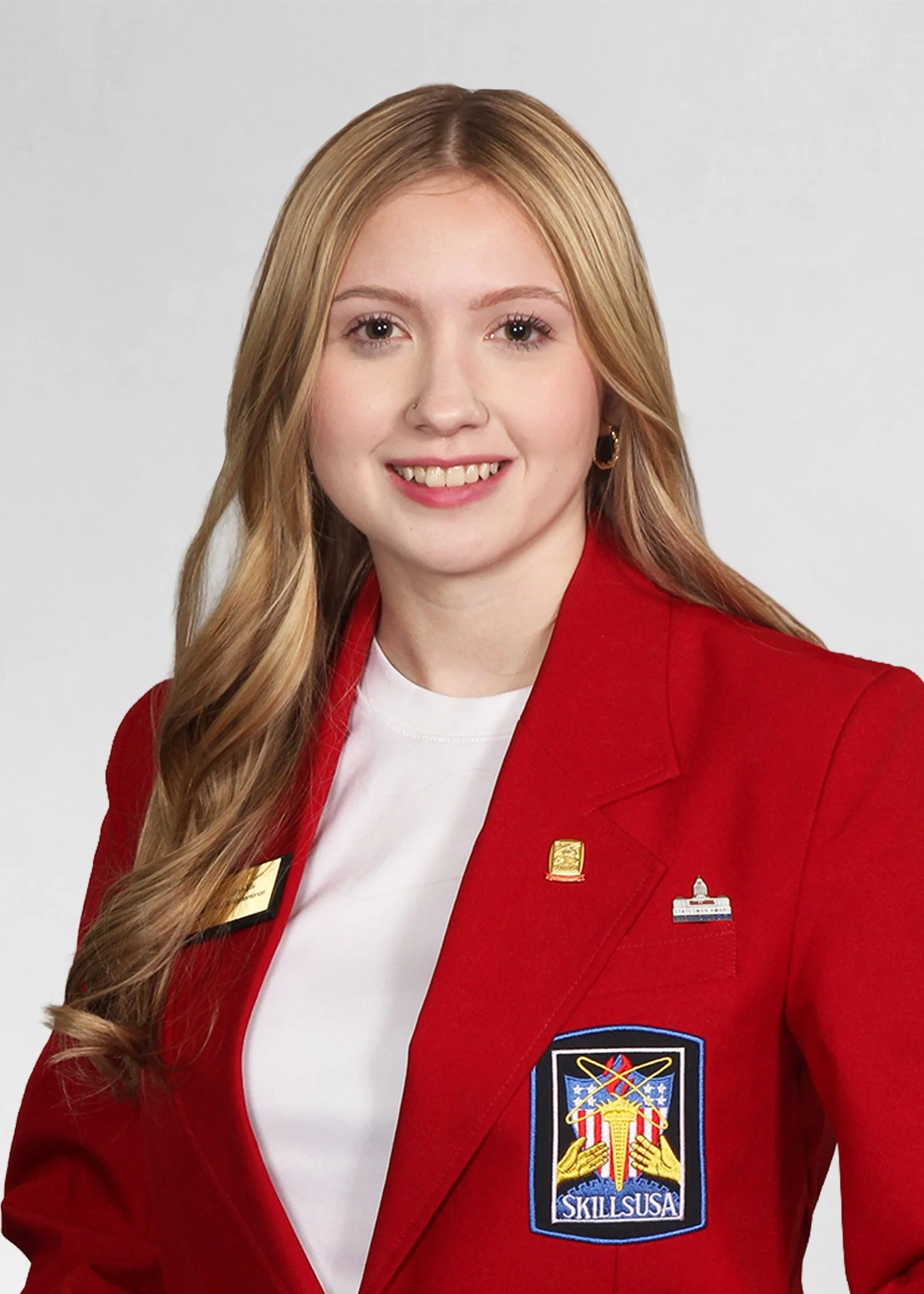 A young woman smiling outdoors, wearing a navy blue jacket with 'SkillsUSA Massachusetts' and 'State Officer' embroidered on it, with a cityscape and green trees in the background.