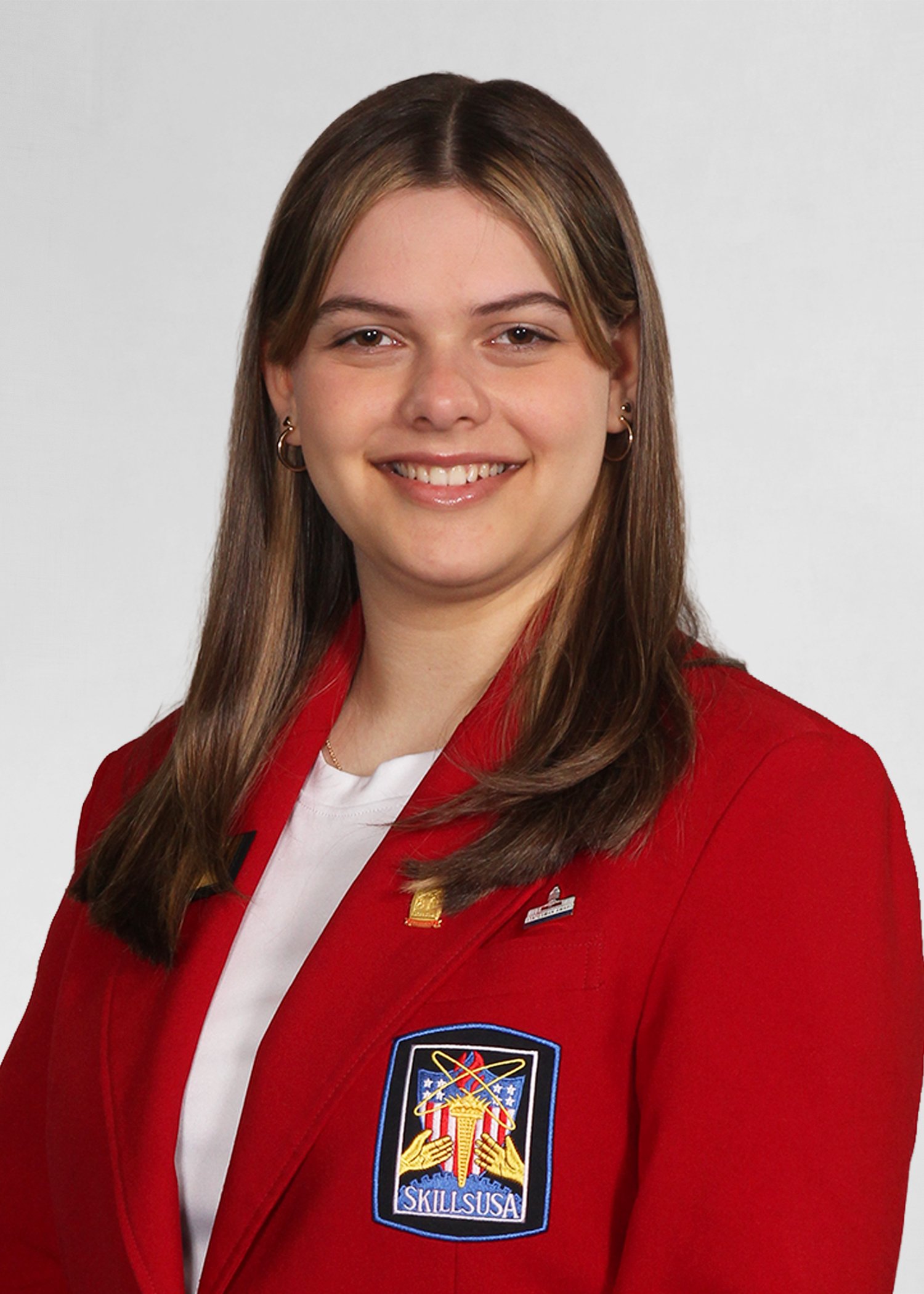 Young woman smiling outdoors, wearing a navy SkillsUSA Massachusetts jacket with 'State Officer' embroidered on it, in a city park setting.