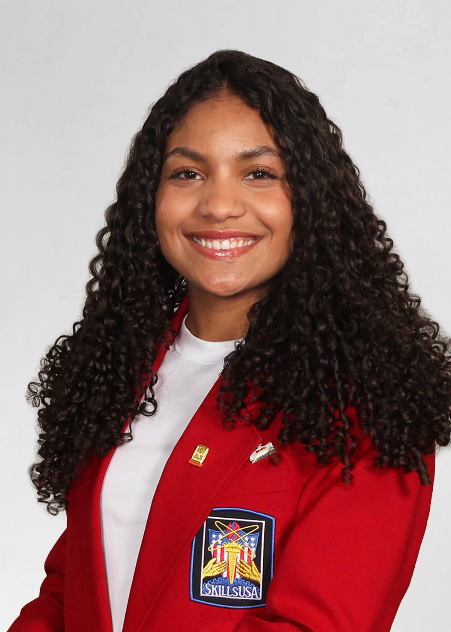 A woman smiling outdoors, wearing a navy blue SkillsUSA Massachusetts jacket with the words 'State Officer' embroidered on it. She has curly black hair, gold hoop earrings, a gold cross necklace, and a gold 'Love' necklace.