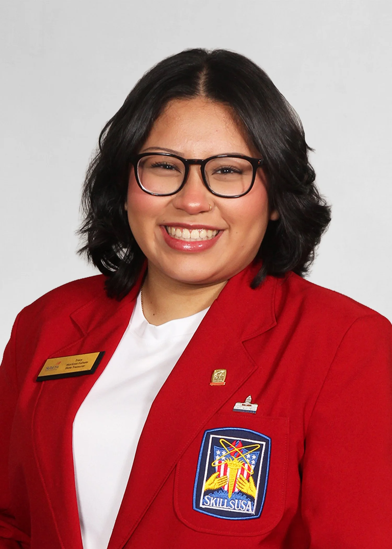 A woman with glasses and dark hair, smiling confidently outdoors, wearing a navy blue jacket with 'SkillsUSA Massachusetts' and 'State Officer' embroidered on it.