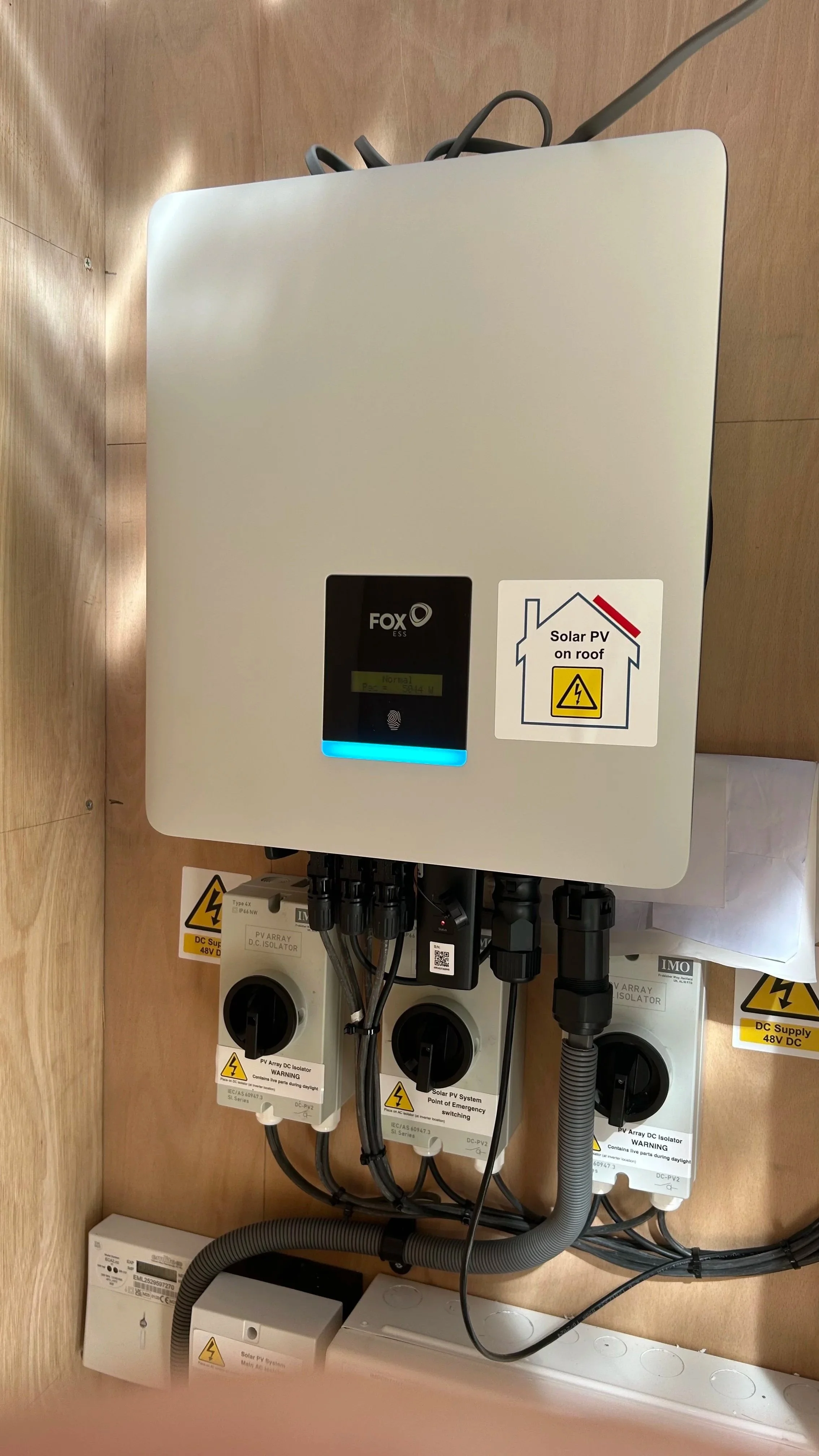 Photovoltaic (PV) system components installed on a wooden wall, including an inverter, circuit breakers, and wiring, with warning and informational labels about solar power on the roof.