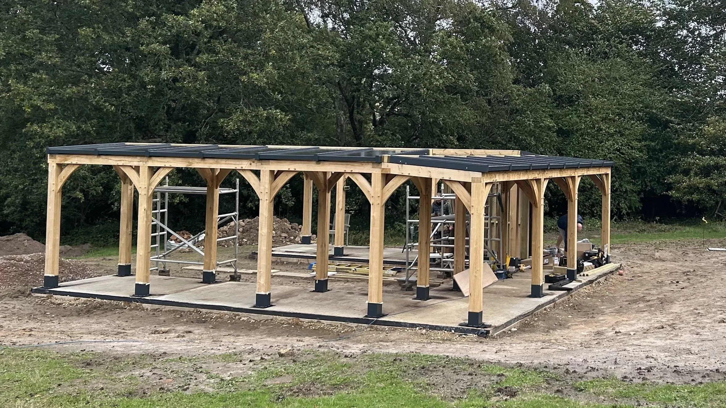Under construction wooden pergola with black metal roofing, surrounded by scaffolding and tools on a concrete slab in an outdoor area with trees in the background.