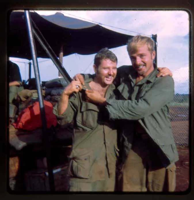 Two smiling men in military uniforms sharing a friendly moment outdoors near a small aircraft.