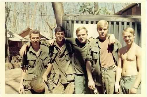 Five young men standing outdoors, some in military-style clothing, one shirtless, posing with arms around each other's shoulders.