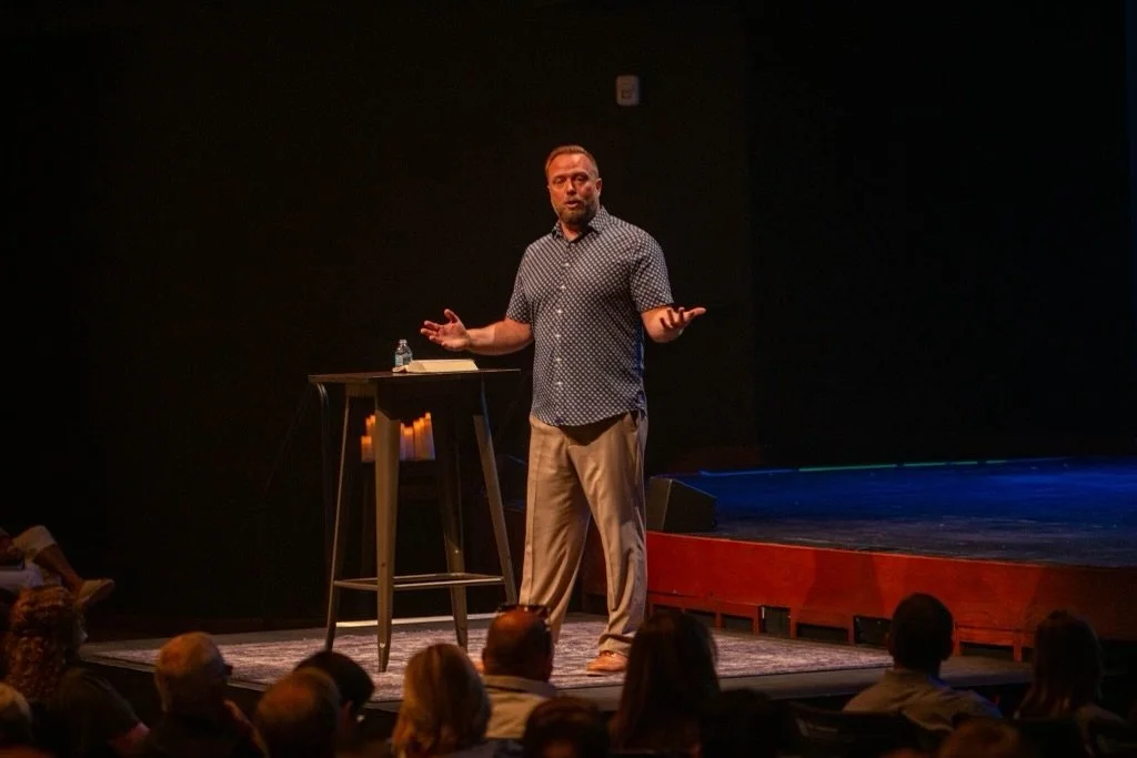 Man giving a presentation on stage with a small round table, audience watching, dark background, stage lighting.