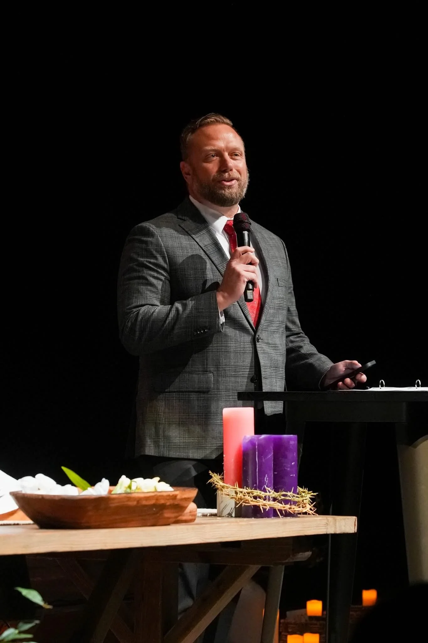 A man in a checked suit is speaking into a microphone at a presentation or event, standing behind a black lectern with candles and decorative items on a table in the foreground.