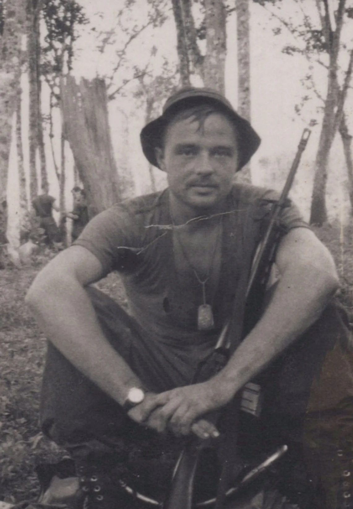 A black and white photo of a man sitting outdoors in a forest, holding a rifle. He is wearing a hat, a wristwatch, and a dog tag necklace. In the background, two other people are seen in the woods.