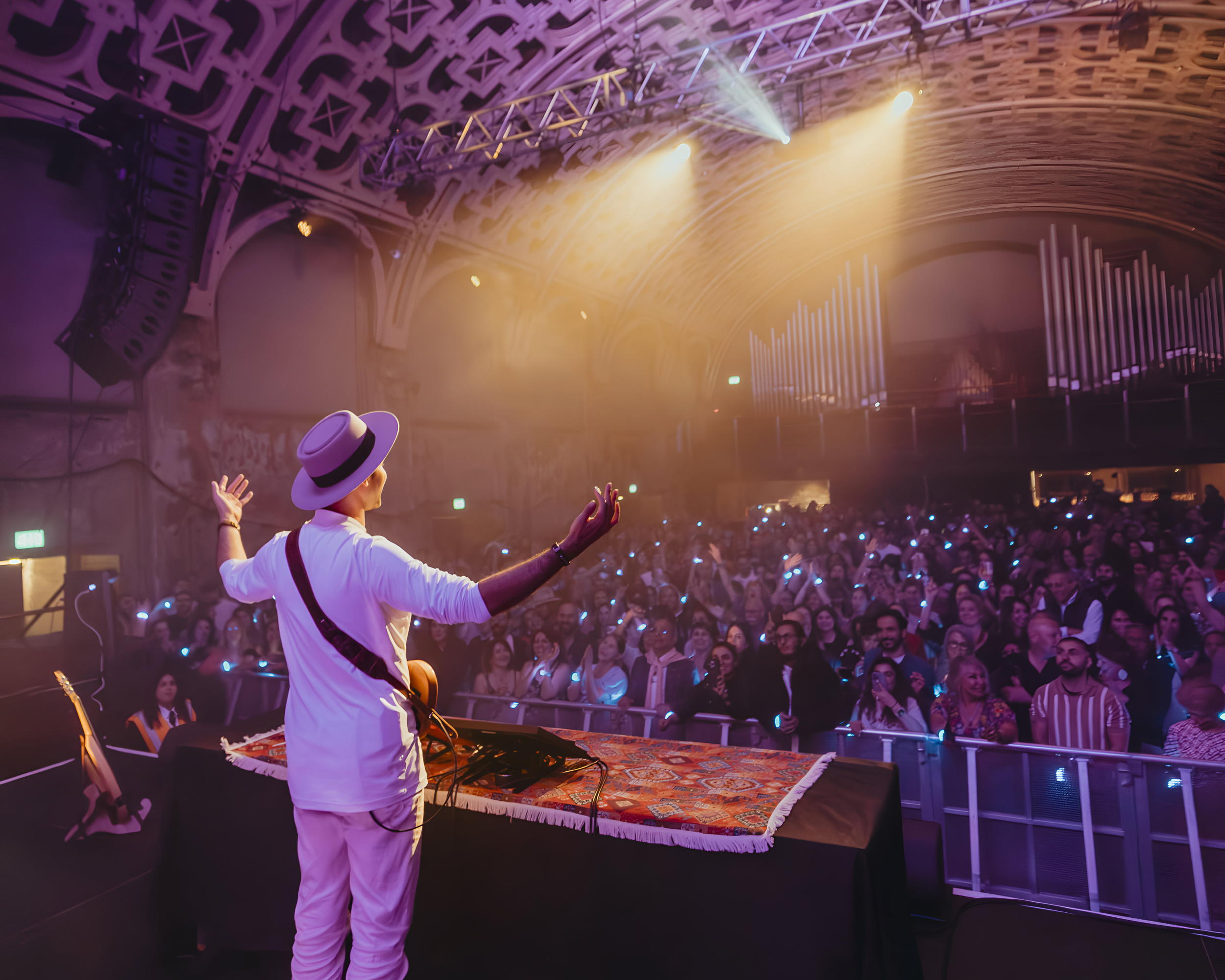 Performer on stage with arms raised, facing large audience in concert hall with vibrant lighting.