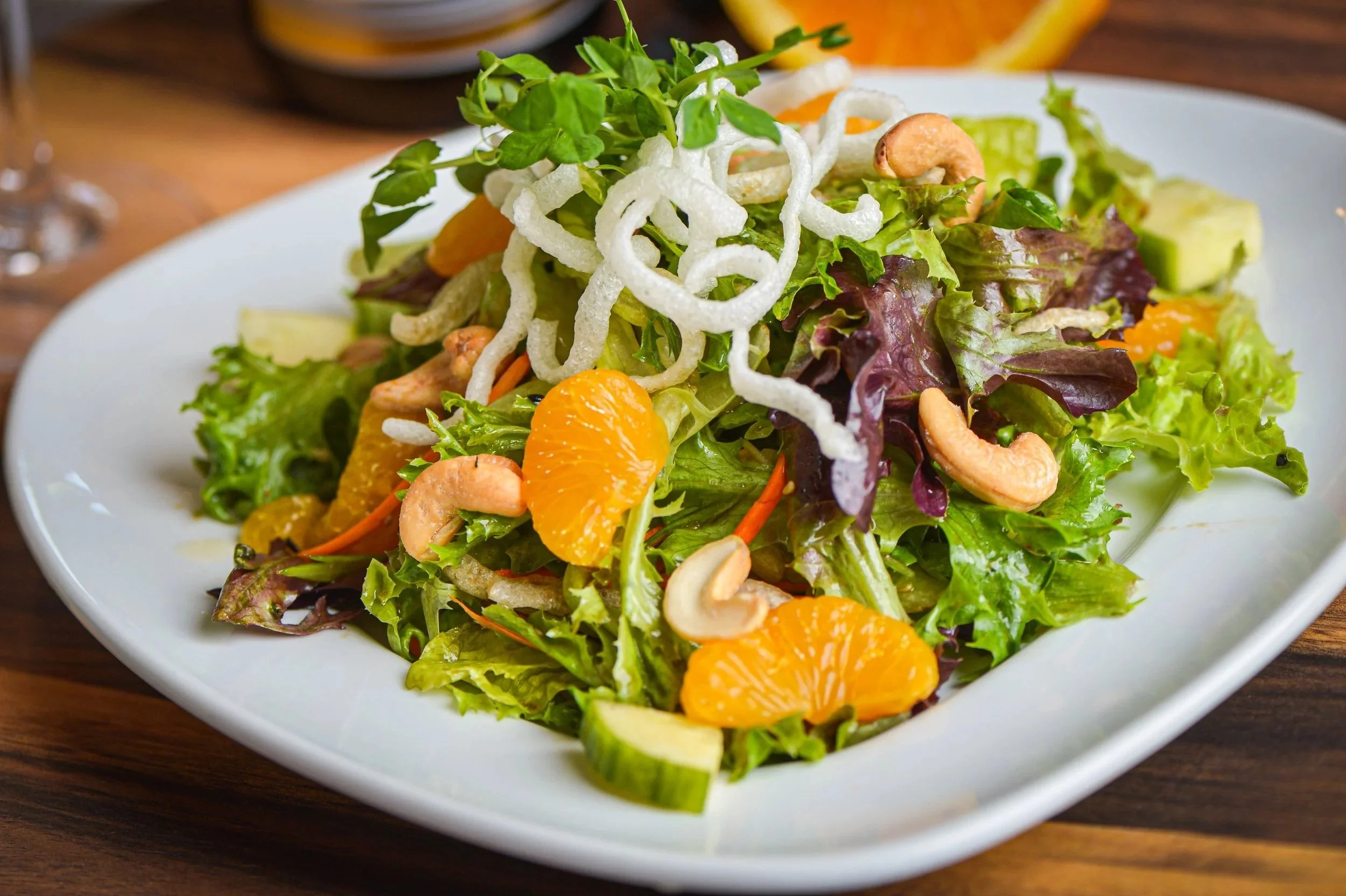 A fresh mixed green salad with carrots, avocado, tangerine segments, cashews, rice crackers, and microgreens on a white plate.
