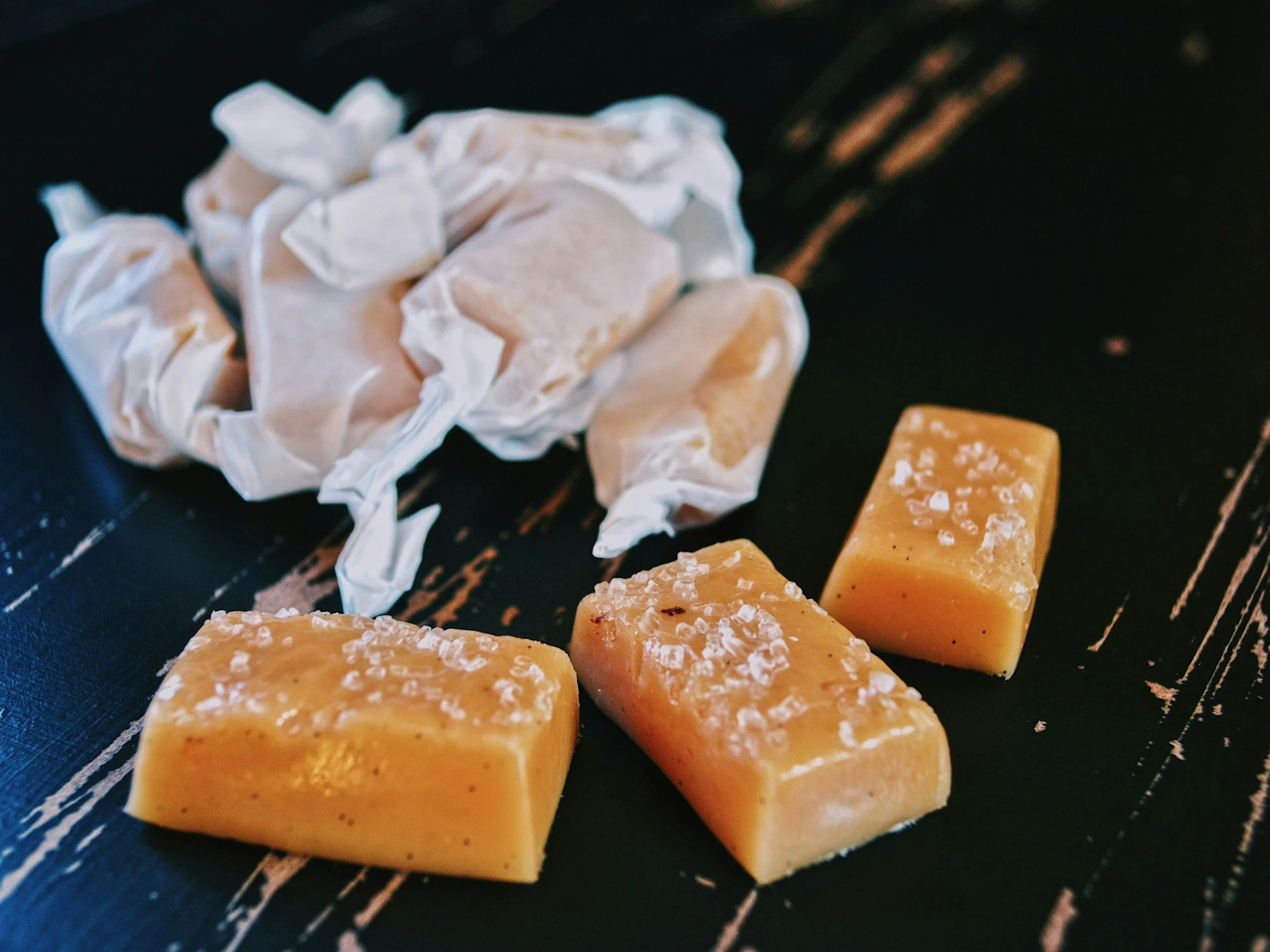 Four blocks of yellowish cheese with salt crystals on top and a crumpled piece of parchment paper on a dark wooden surface.