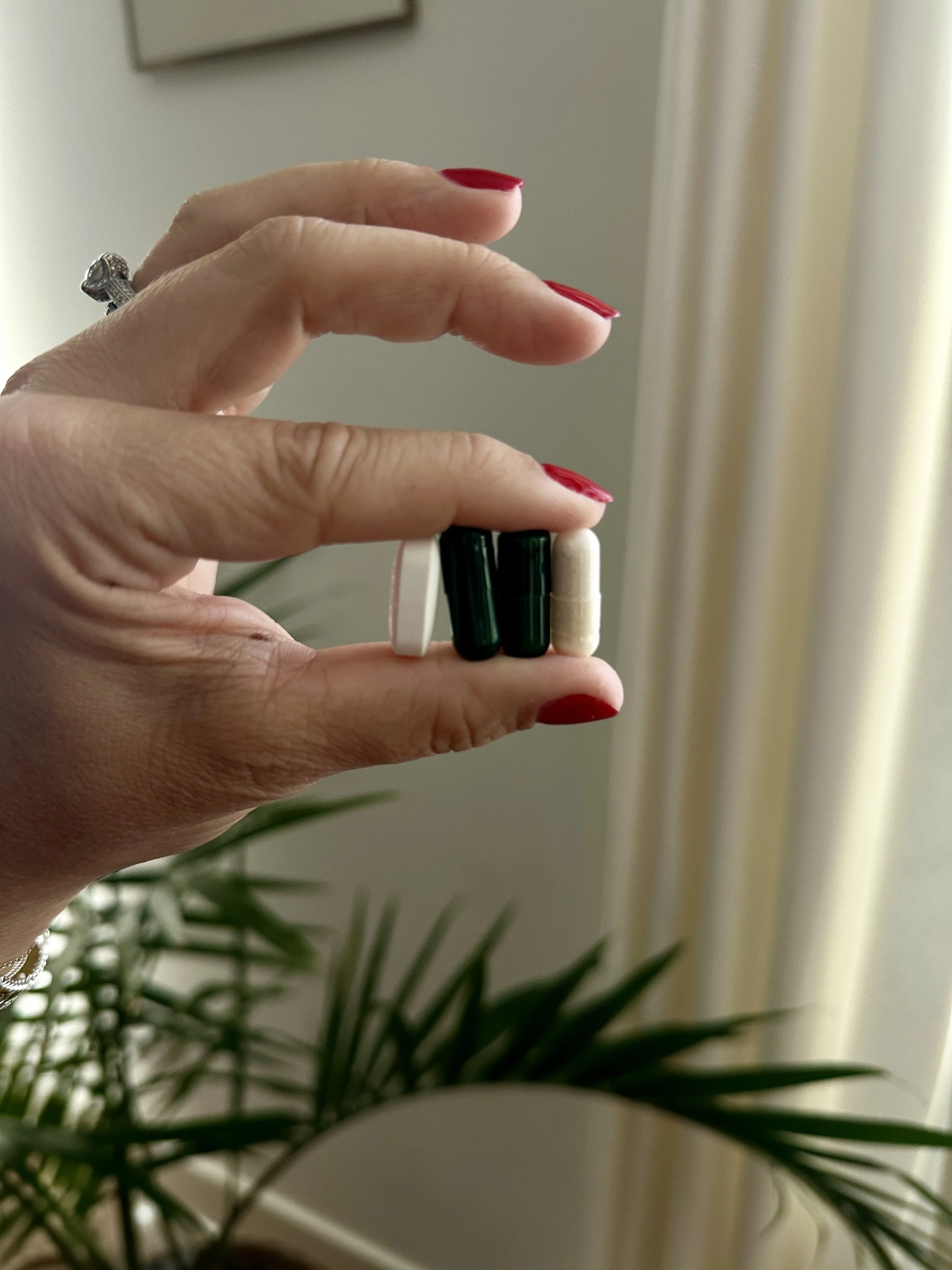 Hand holding two green capsules and two white pills, with red painted nails and a ring on the ring finger.