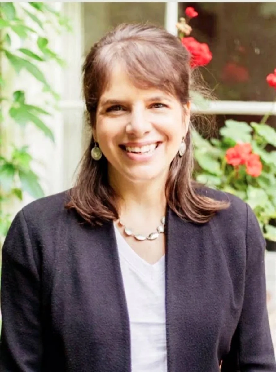 A woman smiling outdoors, wearing a black blazer, white shirt, pearl jewelry, with red flowers and green foliage in the background.