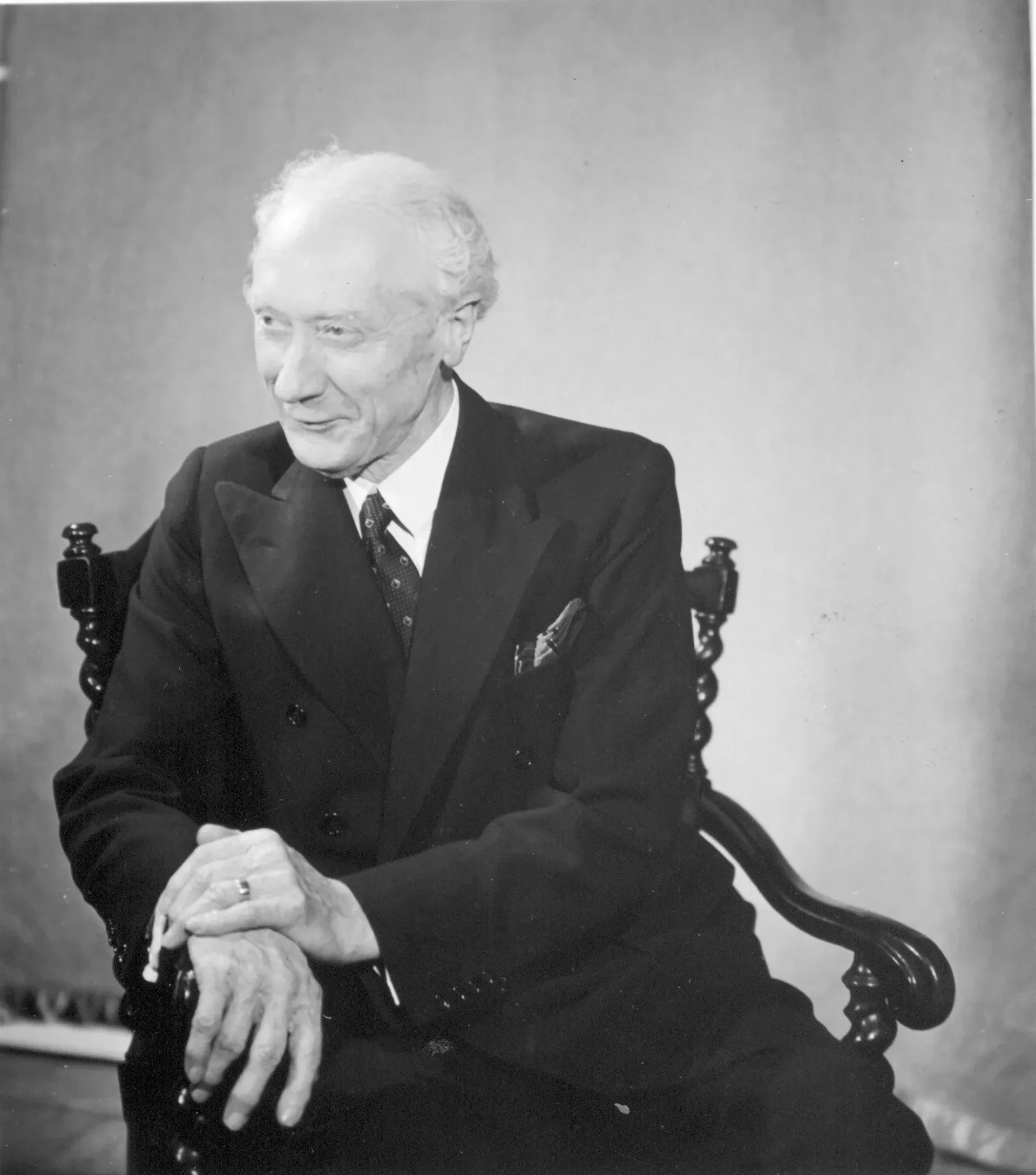 Black and white photograph of an elderly man with wavy white hair, wearing a dark suit, white shirt, and patterned tie, sitting in a wooden chair. His hands are clasped together over a loaf of bread, and he is smiling or speaking softly.