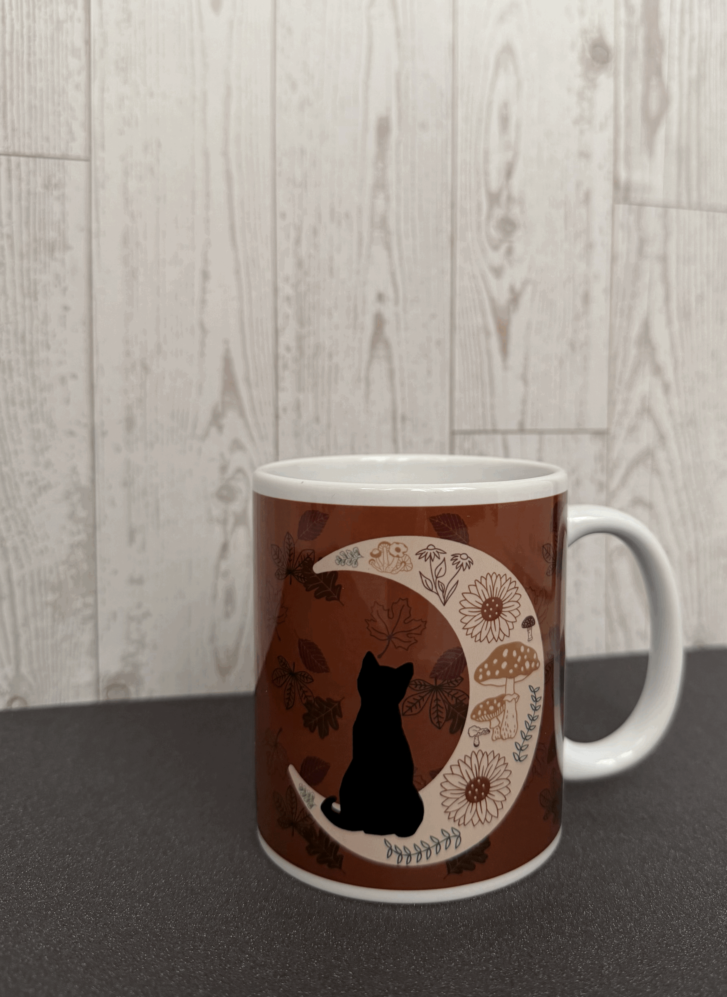 autumn themed coffee mug featuring a black cat watching falling leaves wall sitting on a crescent moon, which is decorated with mushrooms, fall flora, and ferns.gif
