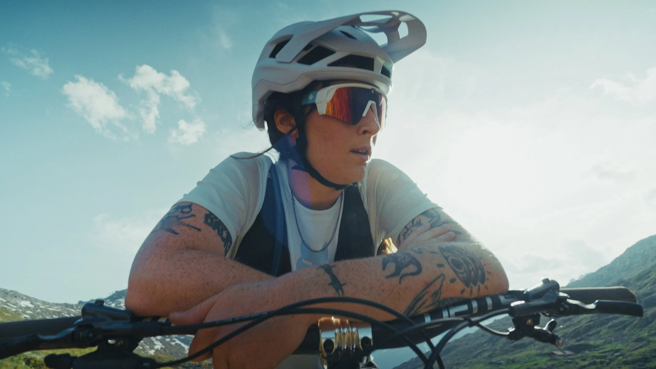 Etalonnage de Kevin Rocha. Une femme portant un casque de vélo, des lunettes de soleil et un maillot blanc, tenant un vélo dans un paysage montagneux ensoleillé.