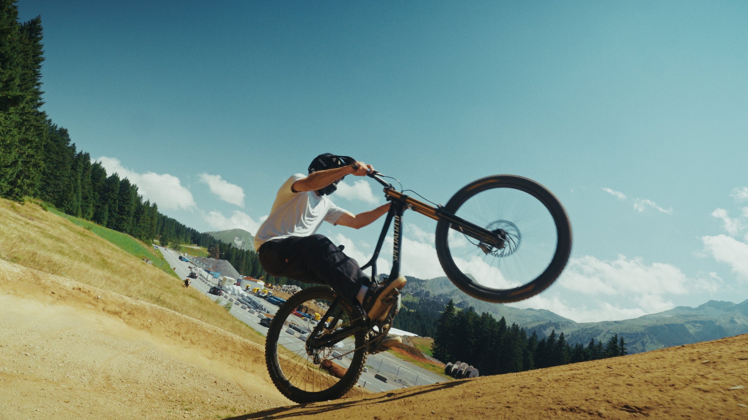 Etalonnage de Kevin Rocha. Un cycliste en plein saut sur une piste de vélo en plein air, montagnes et forêt en arrière-plan, ciel bleu.