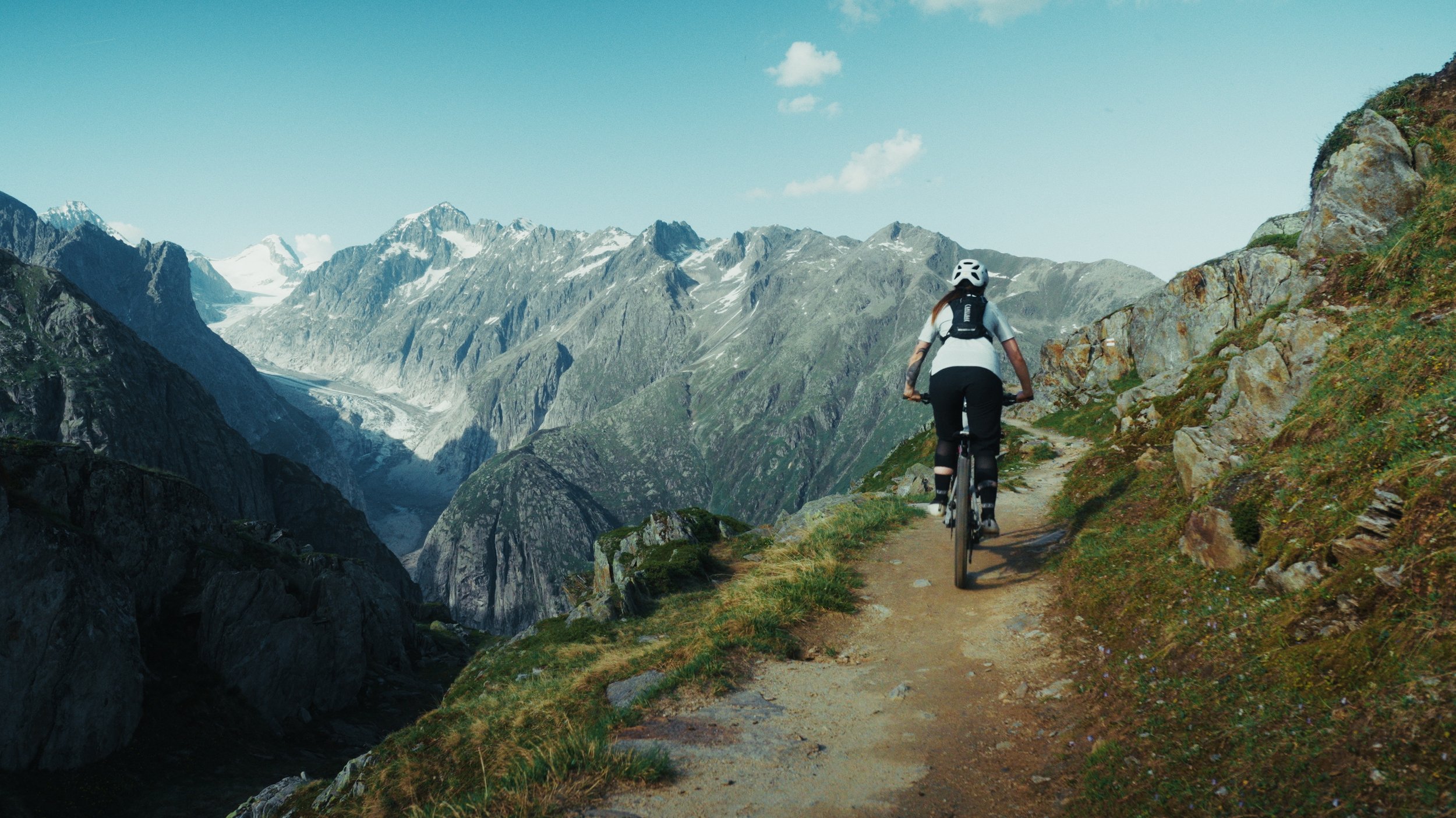 Etalonnage de Kevin Rocha. Une personne fait du cyclisme sur un sentier en montagne avec des sommets enneigés en arrière-plan.