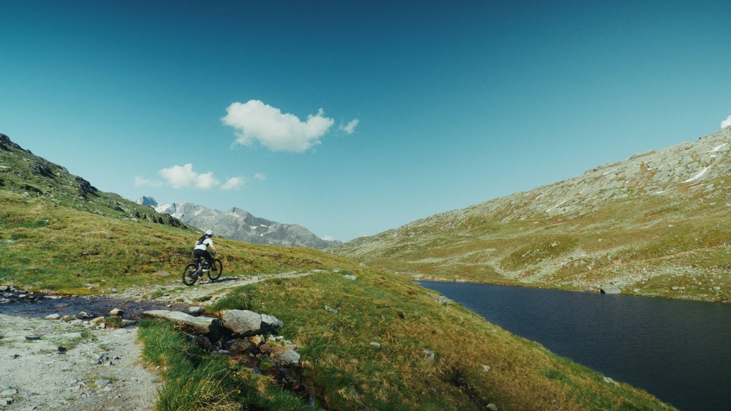 Etalonnage de Kevin Rocha. Un cycliste pédale sur un sentier dans un paysage montagneux avec un lac à droite, des montagnes enneigées en arrière-plan, et un ciel bleu avec quelques nuages.
