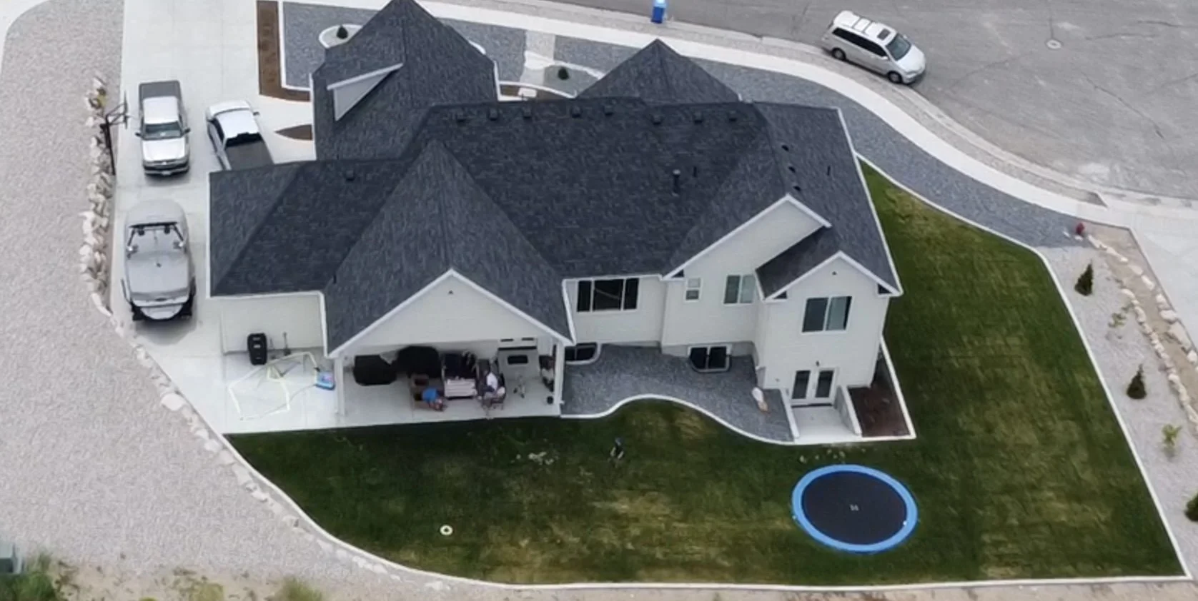 Aerial view of a large modern house with a dark gray roof, surrounded by a green lawn, a white gravel driveway, and a small blue trampoline in the backyard. Several cars are parked near the house, with some on the driveway and street. There are outdoor seating and a patio area with a canopy and a few children playing.