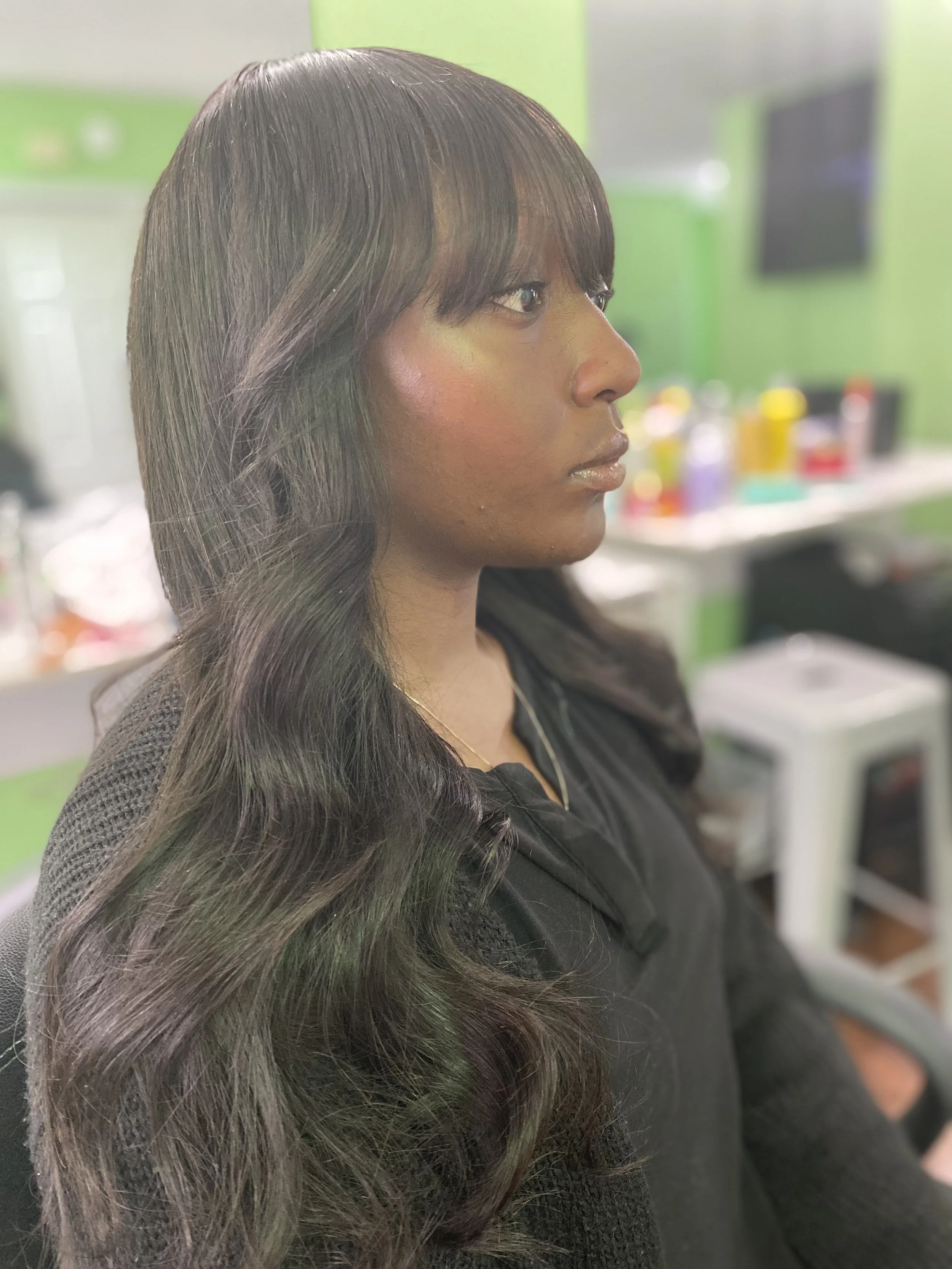 Side profile of a woman with long wavy dark hair and bangs, sitting indoors with a green wall and blurred background of salon or barbershop supplies.