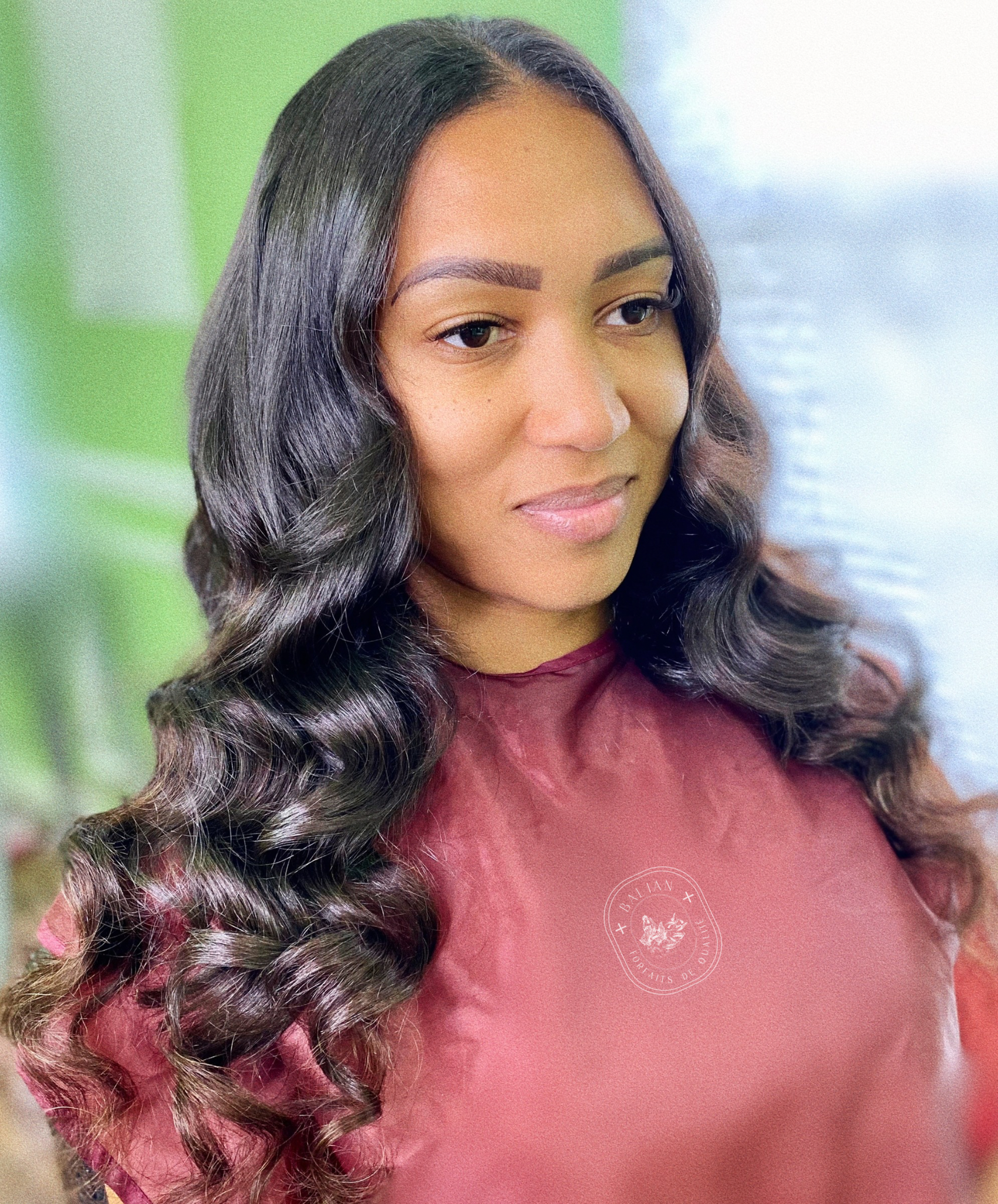 A woman with long, wavy, dark hair and natural makeup, wearing a maroon top with a subtle logo, standing against a background of blurred green and blue tones.