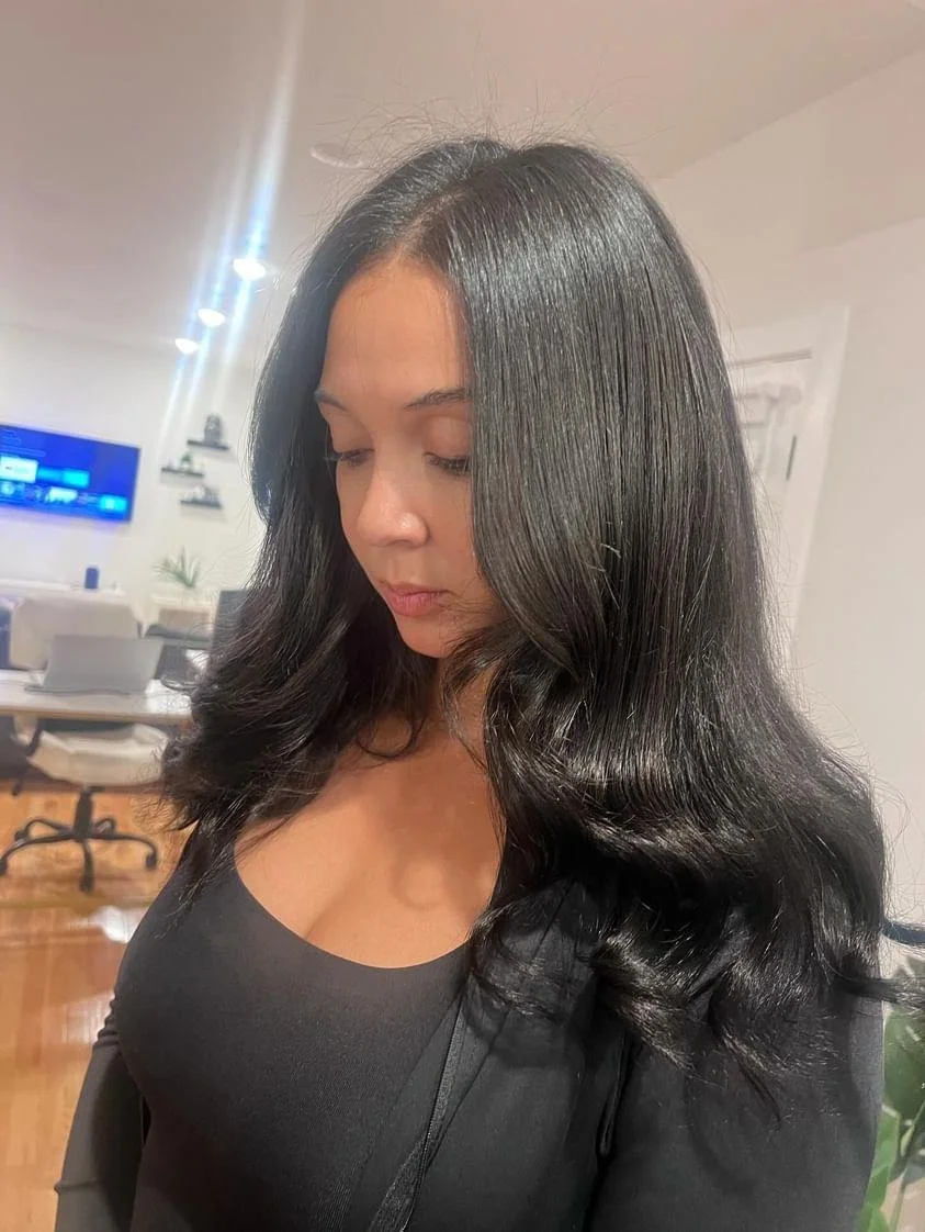 A woman with long, shiny black hair and fair skin standing indoors in a room with white walls and a wooden floor, wearing a black top.