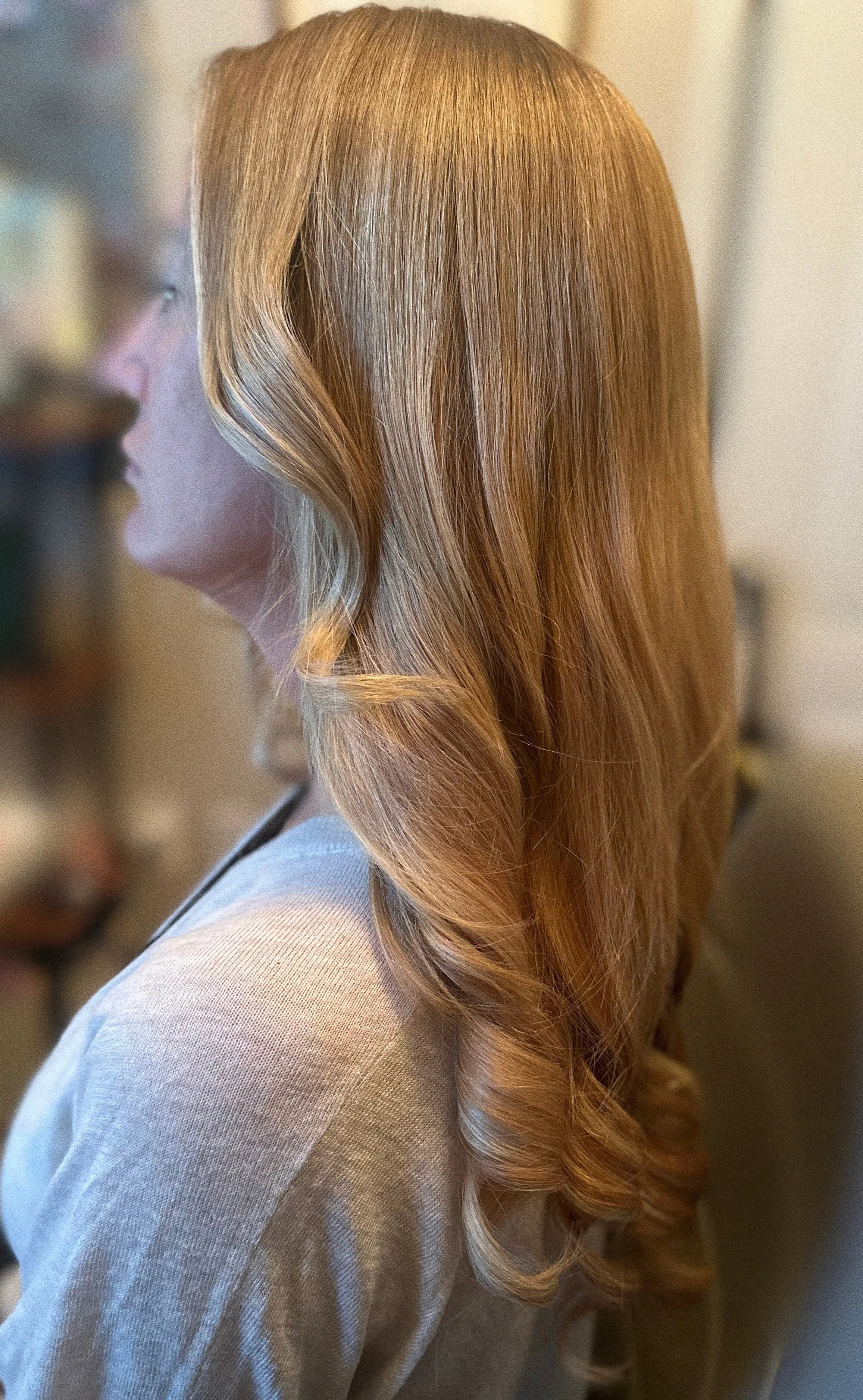 A woman with long, wavy red hair seen in profile from the shoulders up, wearing a light gray top, indoors with blurred background.