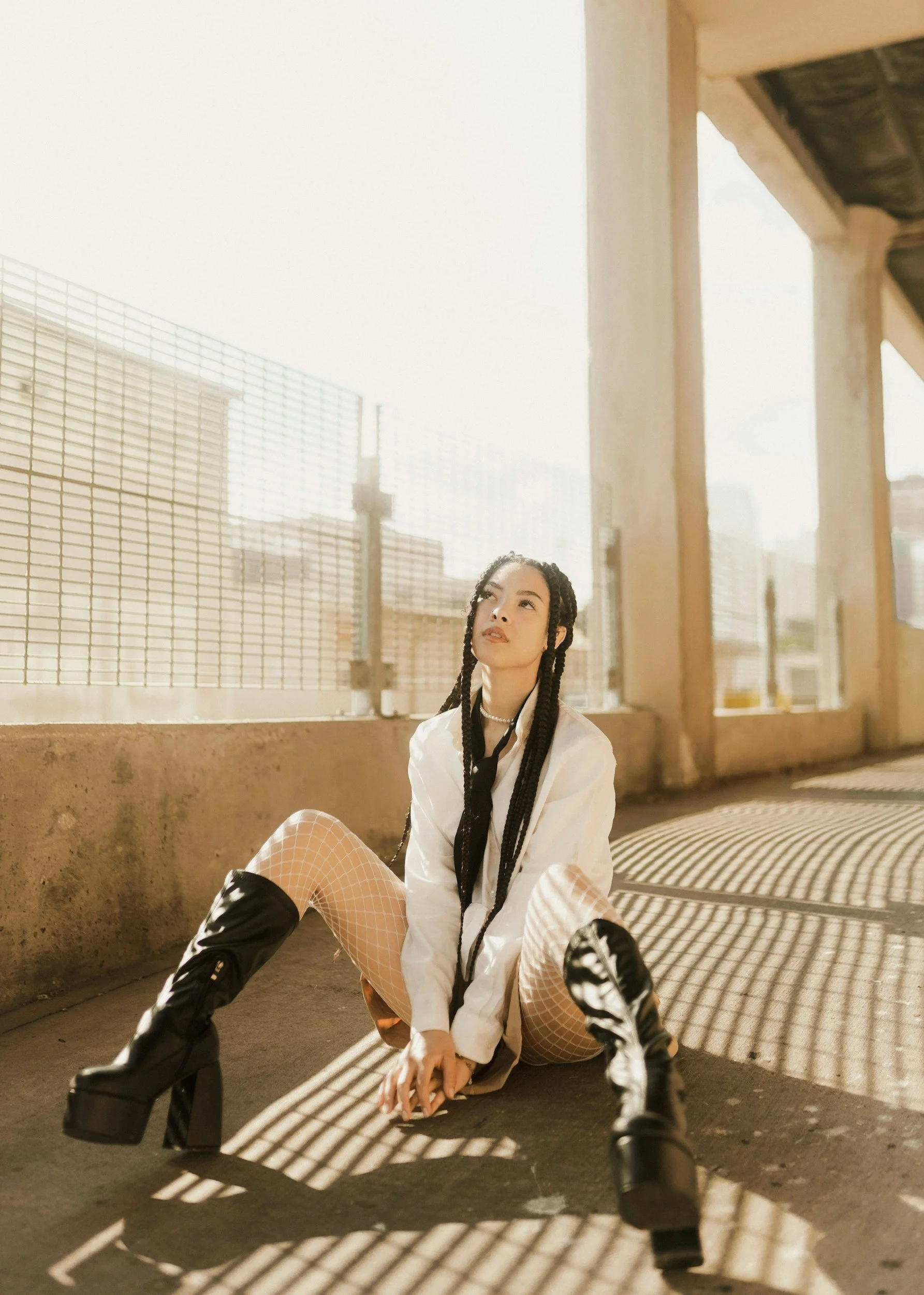 Person with long braids sitting on a walkway under an urban bridge, wearing a white shirt, fishnet stockings, and chunky black boots.
