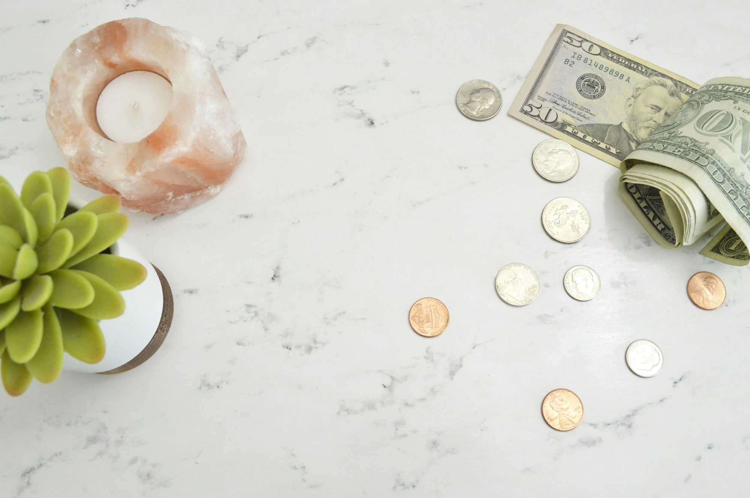 A white table with a small green succulent plant in a white pot, a Himalayan salt candle holder with a white candle, and scattered U.S. currency including a rolled-up $50 bill, a $20 bill, and various coins.