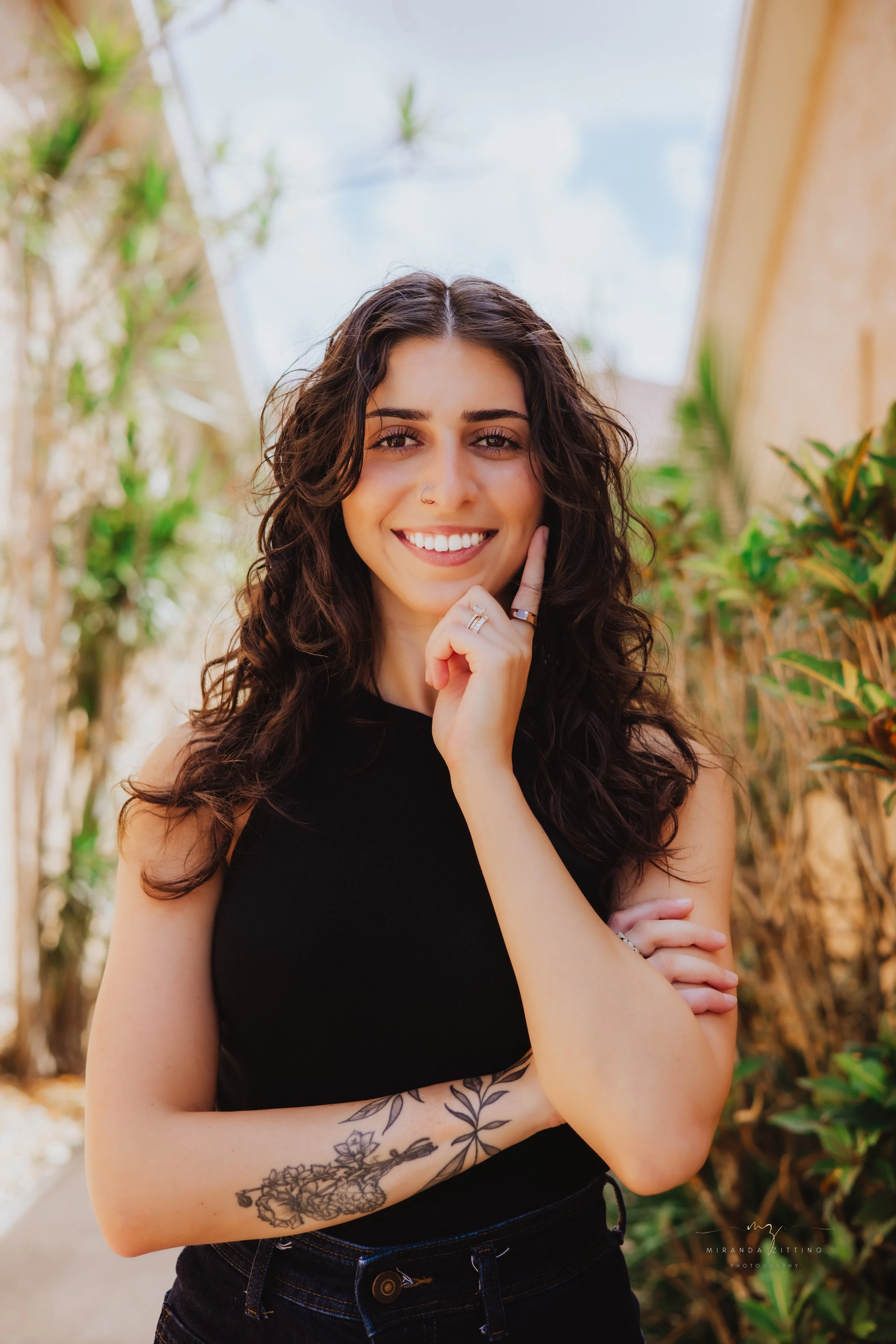 Young woman with curly dark hair, smiling, wearing a black sleeveless top and jeans. She has tattoos on her left arm and multiple rings on her fingers. She is standing outdoors among plants.