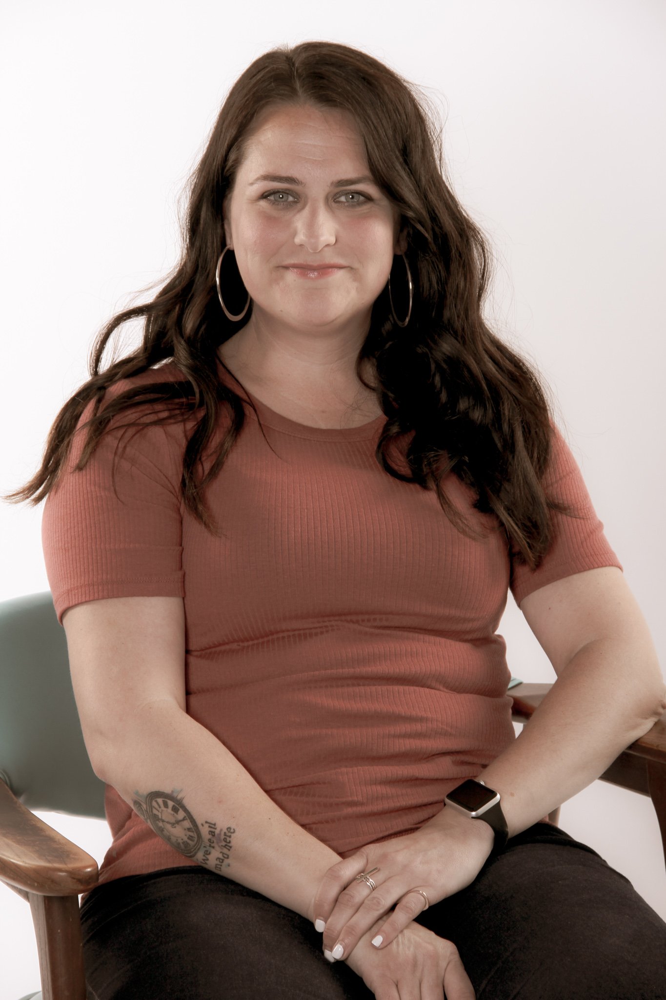 Jennifer Vincent, Therapist with long dark hair sitting on a chair, wearing a rust-colored top and black pants, with a tattoo on their left forearm and a smartwatch on their right wrist.
