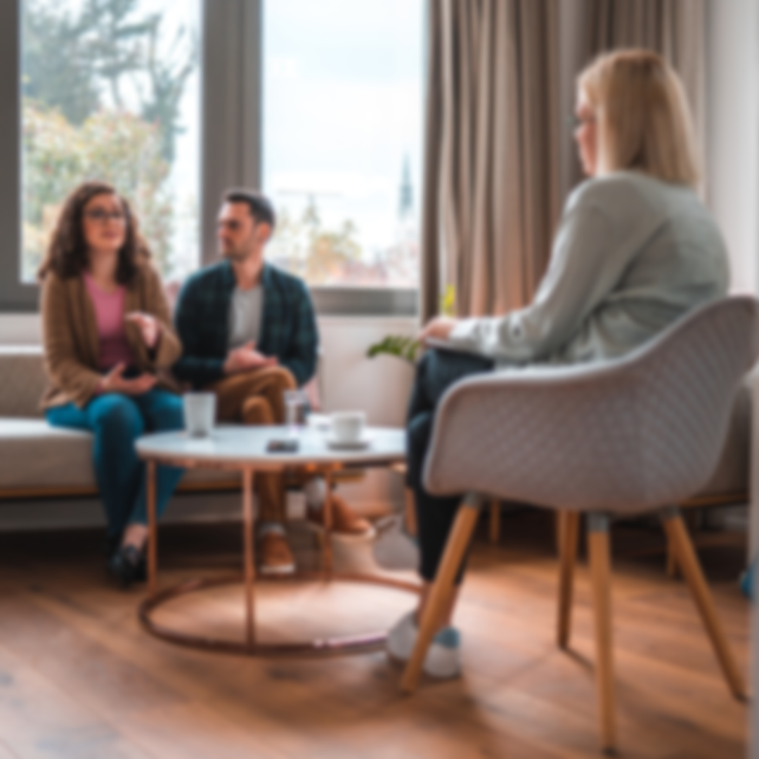 A woman having a therapy session with a couple in a bright living room with large windows and beige curtains.