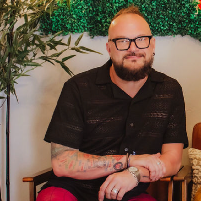 A man with glasses, a beard, and tattoos on his left arm, wearing a black shirt and red pants, sitting in front of a green leafy wall and a potted plant.