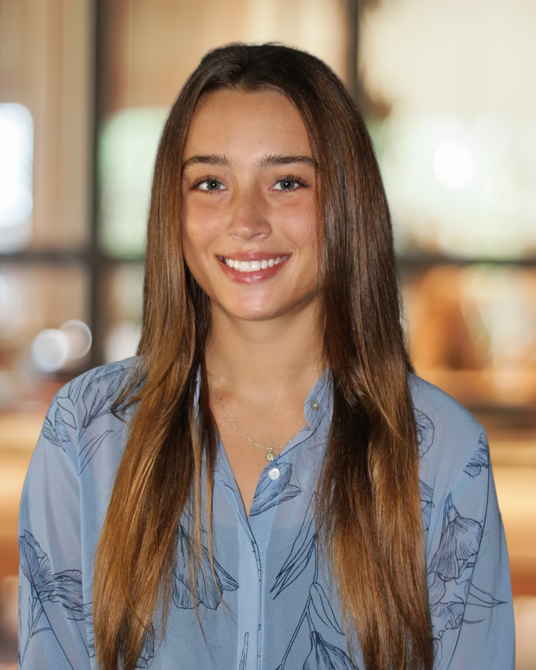Smiling woman with long brown hair, glasses, and a gray sweater sitting outdoors near a green leafy plant and glass window.