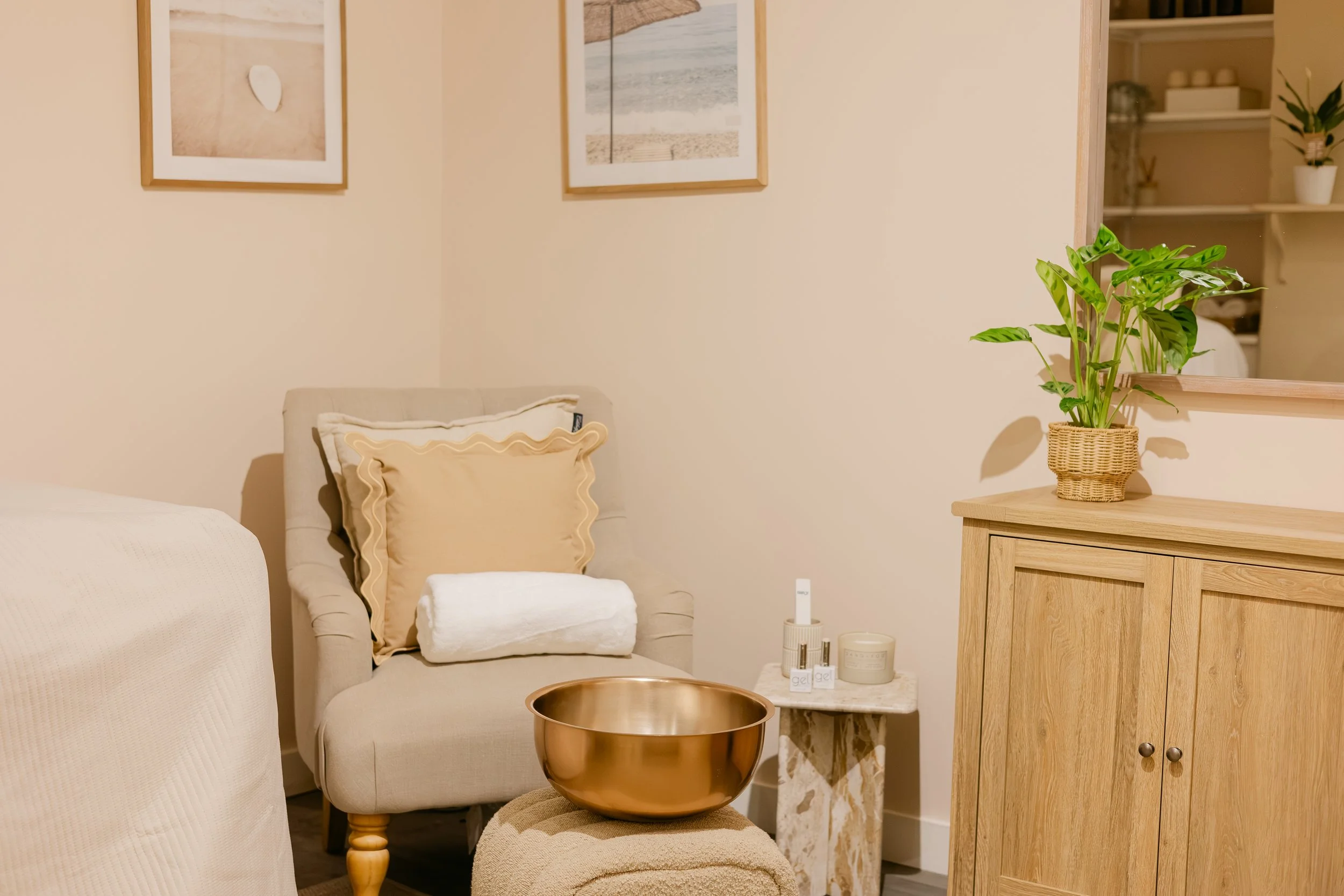Cozy bedroom corner featuring a beige armchair with a cream pillow and a rolled towel. A small marble side table holds skincare products. A large brass bowl sits on a striped ottoman. A wooden dresser with a potted plant is next to a mirror. Two fram