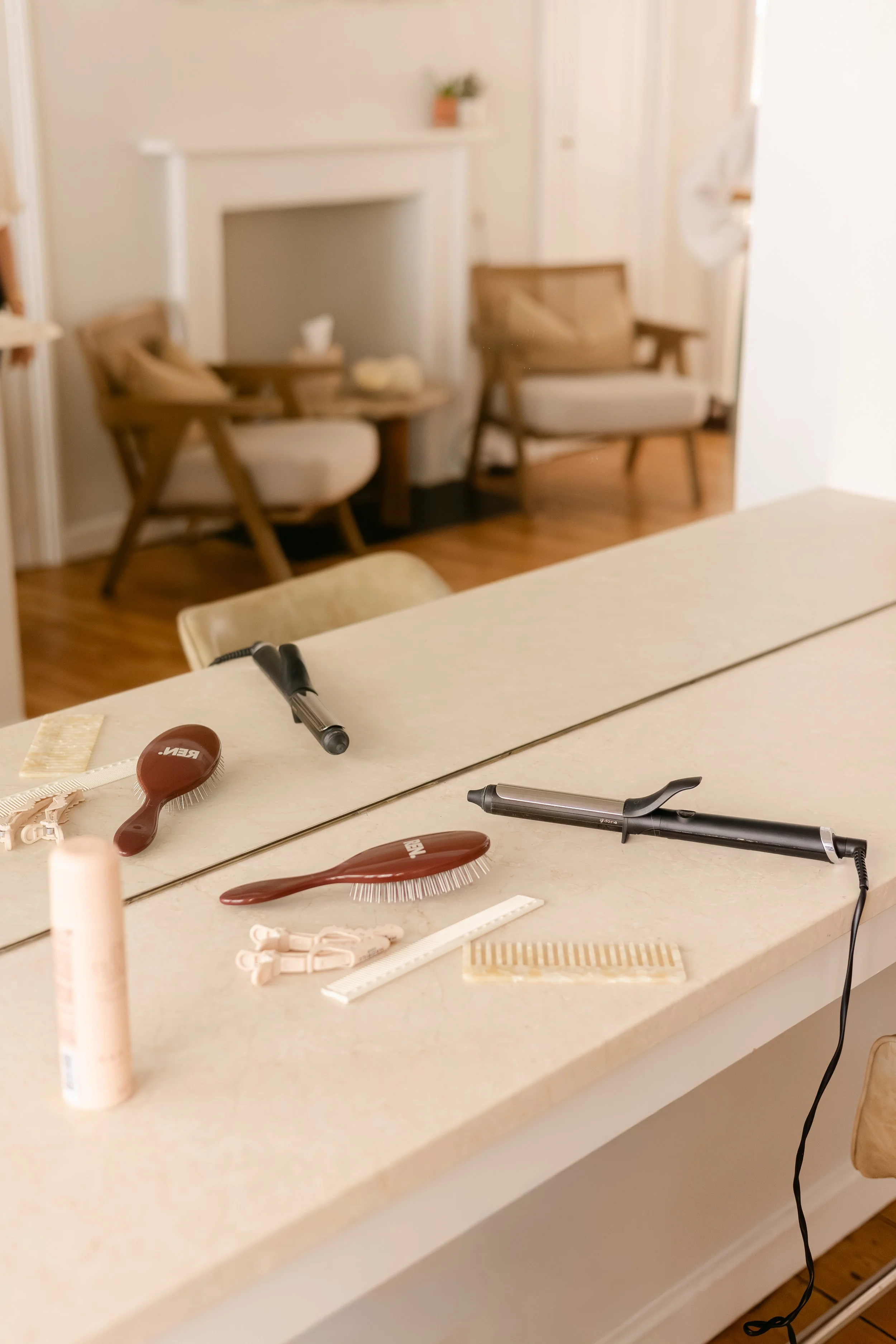 A countertop with various hair styling tools including brushes, a round brush, a comb, and a curling iron, with blurred chairs and a fireplace in the background.