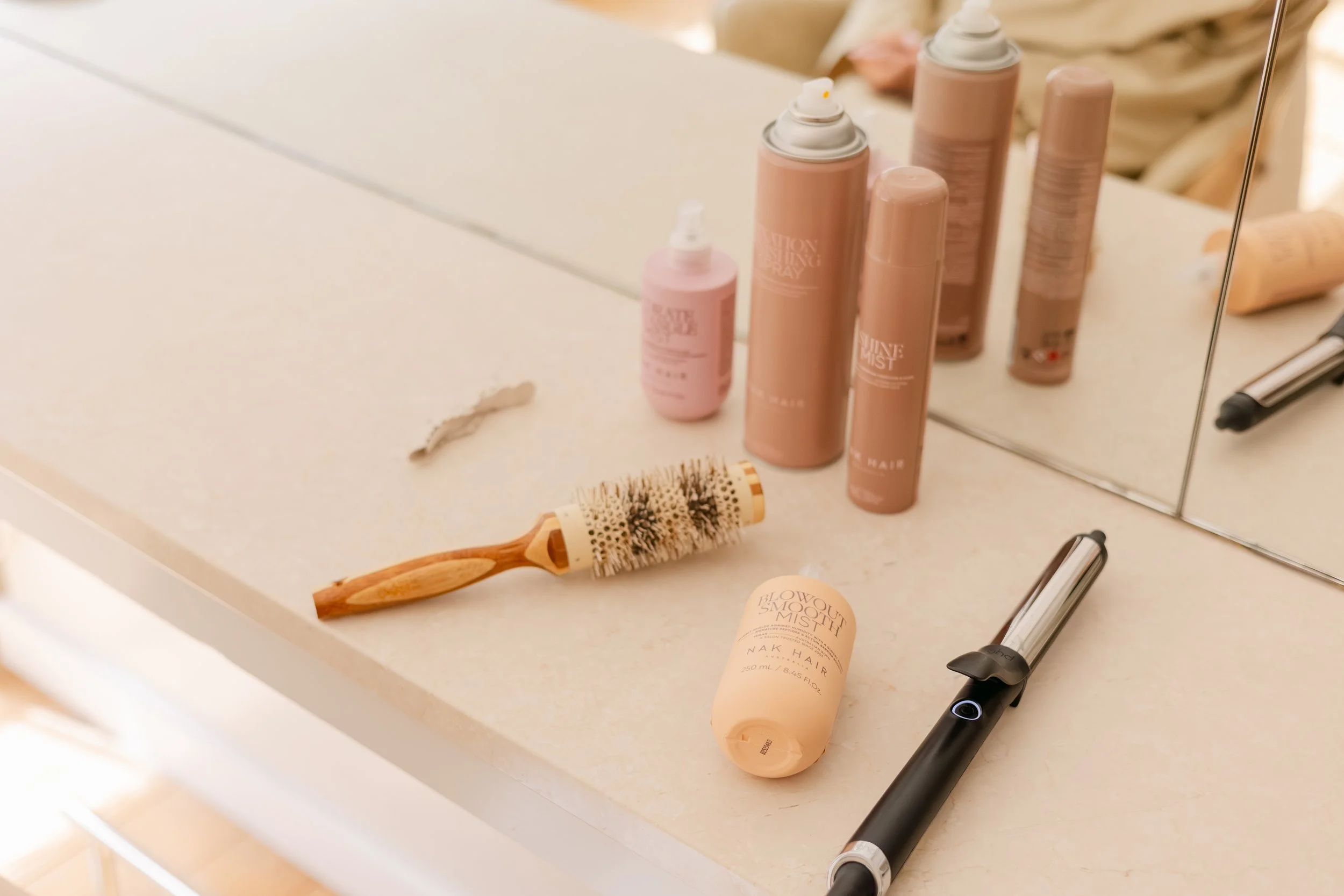 A bathroom countertop with various hair care and beauty products, including spray bottles, a hairbrush, and a tube of cosmetic product, reflected in a mirror.