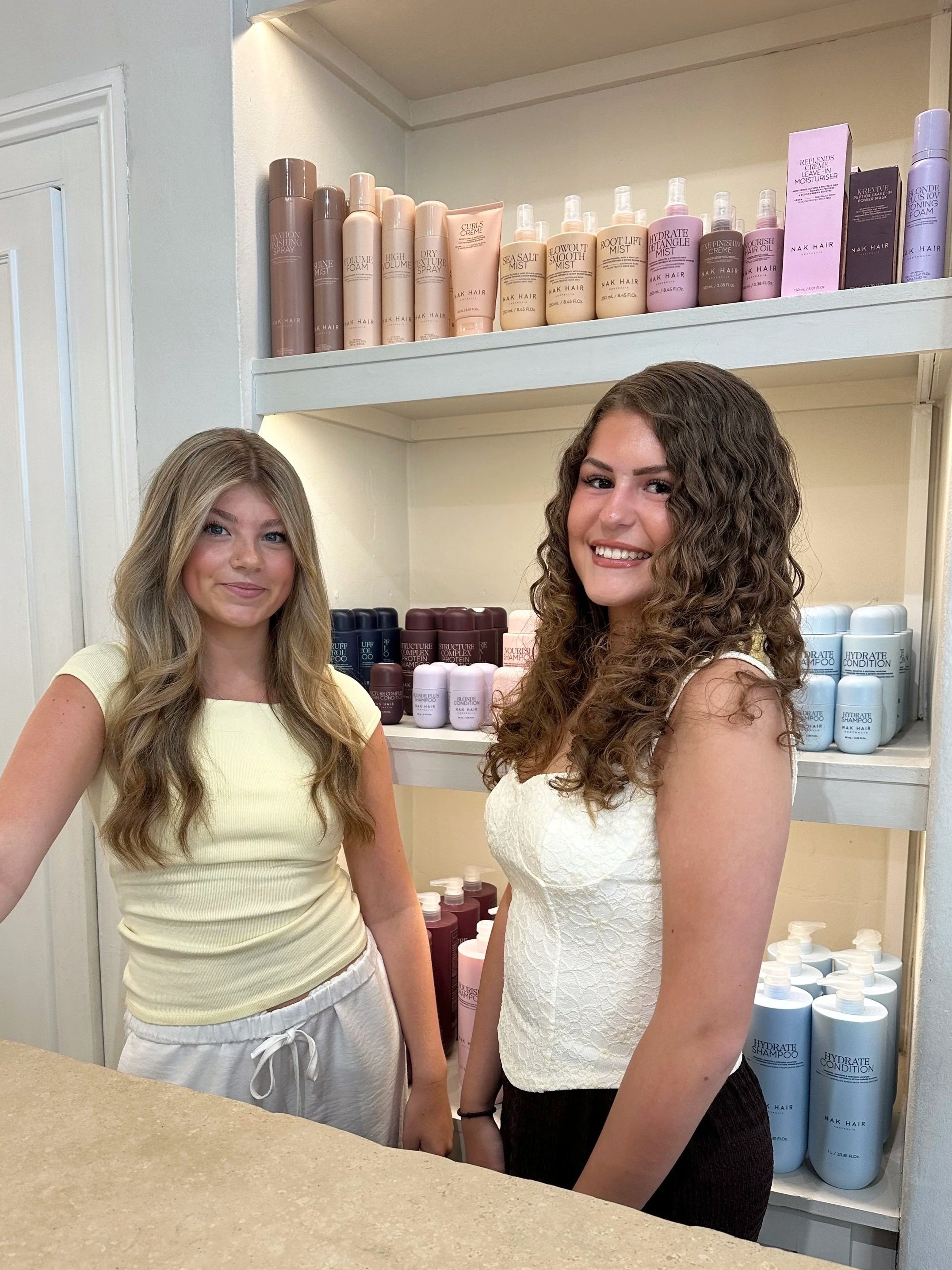 Two 12 Trim Street apprentices, behind them are shelves with various hair care products, including shampoo and conditioner bottles.