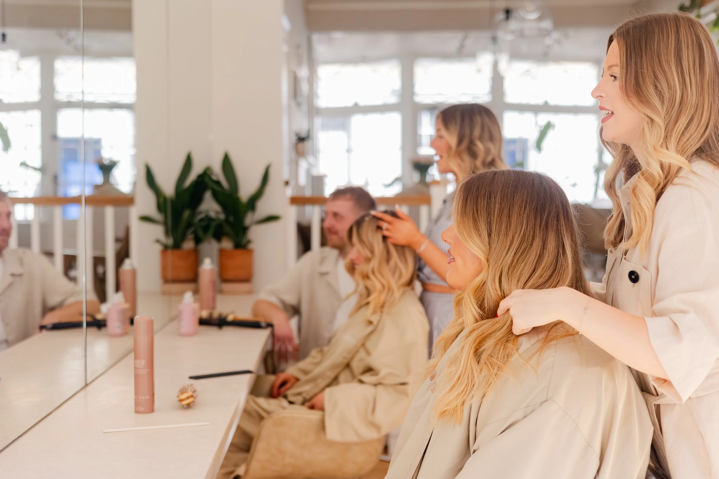 A group of women sitting at a long table in a brightly lit room, with one woman standing and smiling, other women engaged in conversation, and hair styling products on the table.